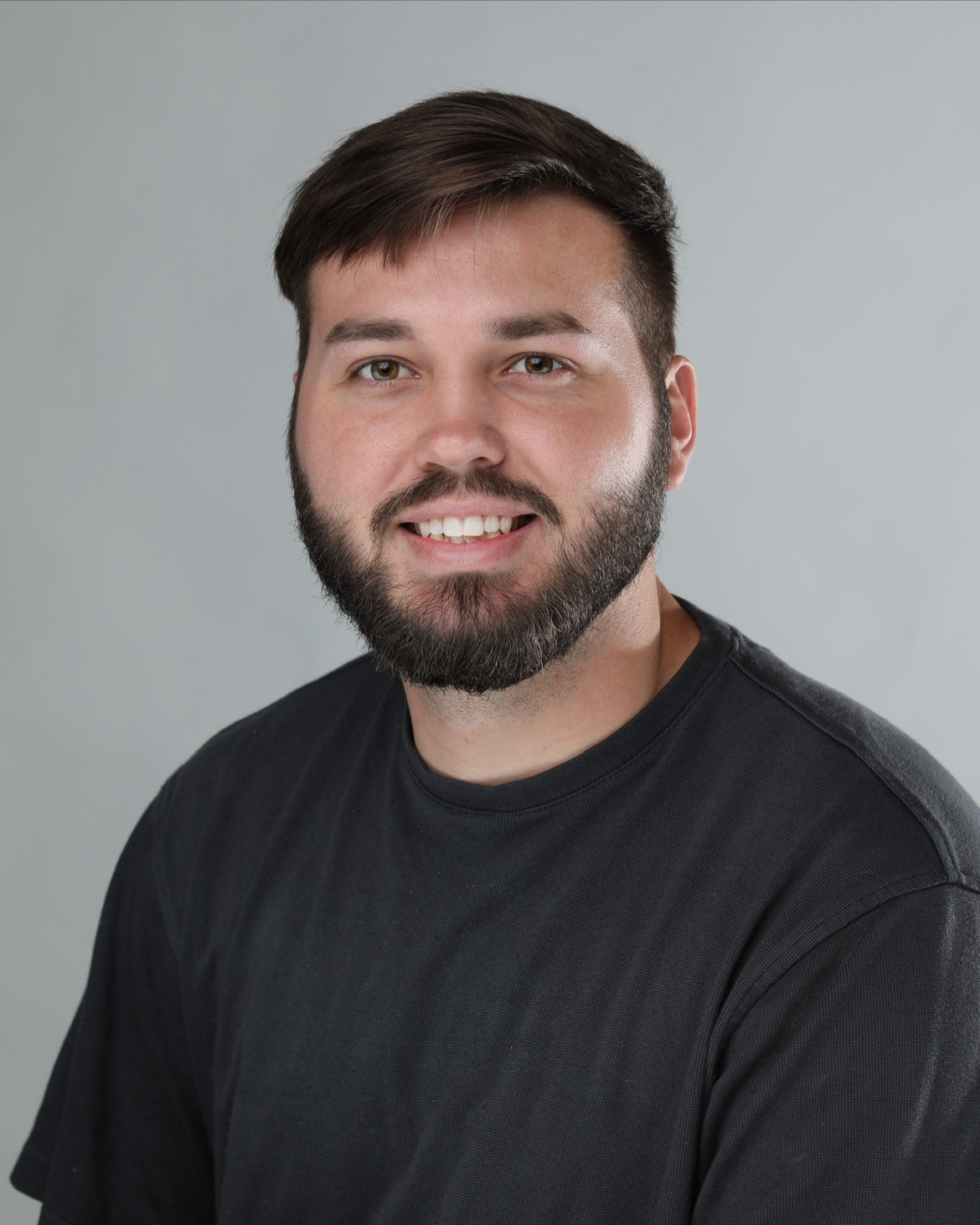 A young man with a beard and short brown hair smiling, wearing a black T-shirt, against a plain gray background.