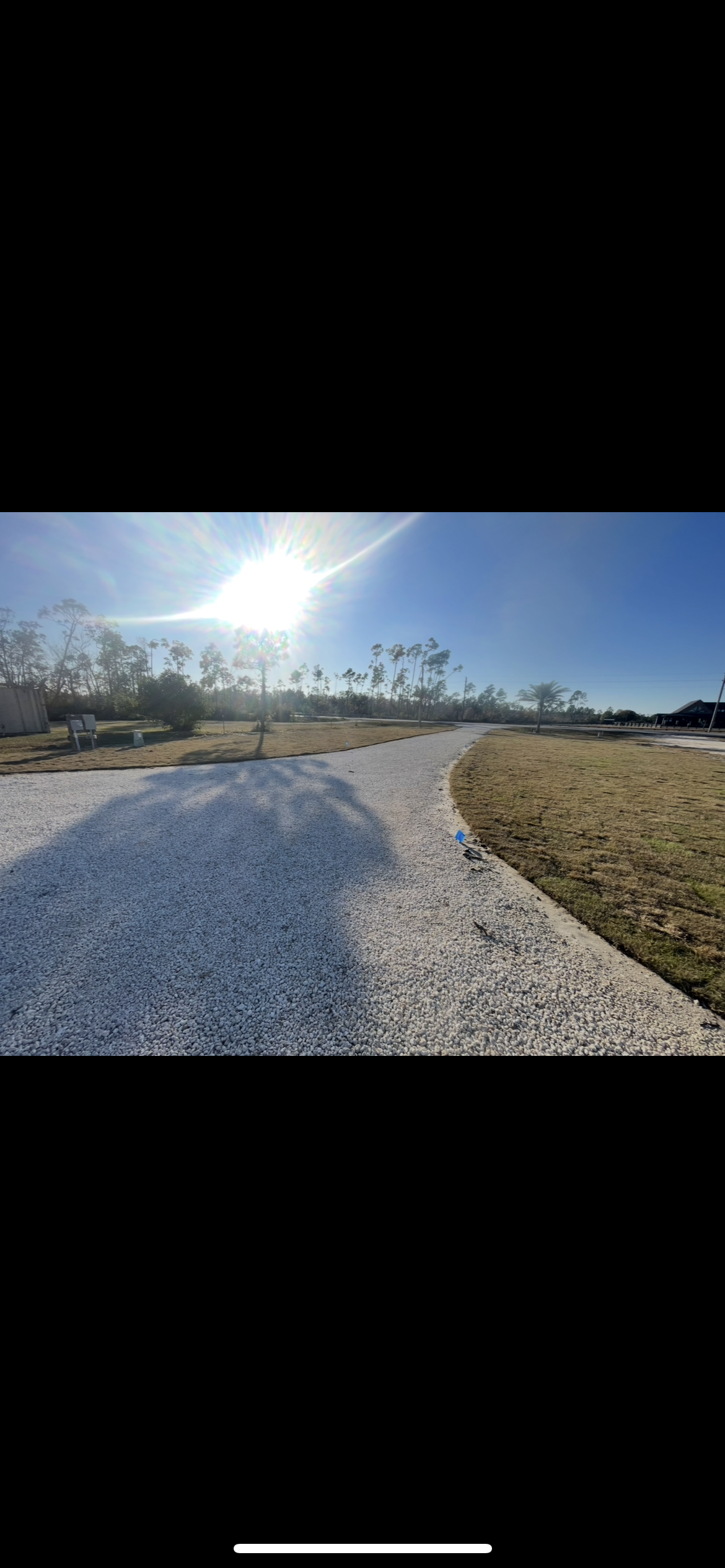 Sun shining brightly over a gravel pathway with shadows of trees, a clear blue sky, and some trees and a building in the background.