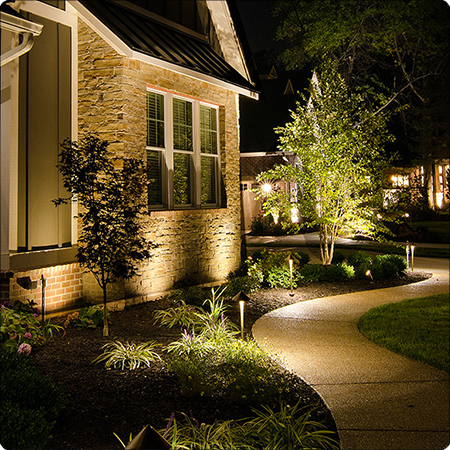 Night view of a house yard with illuminated garden lights, curved pathway, and trees.