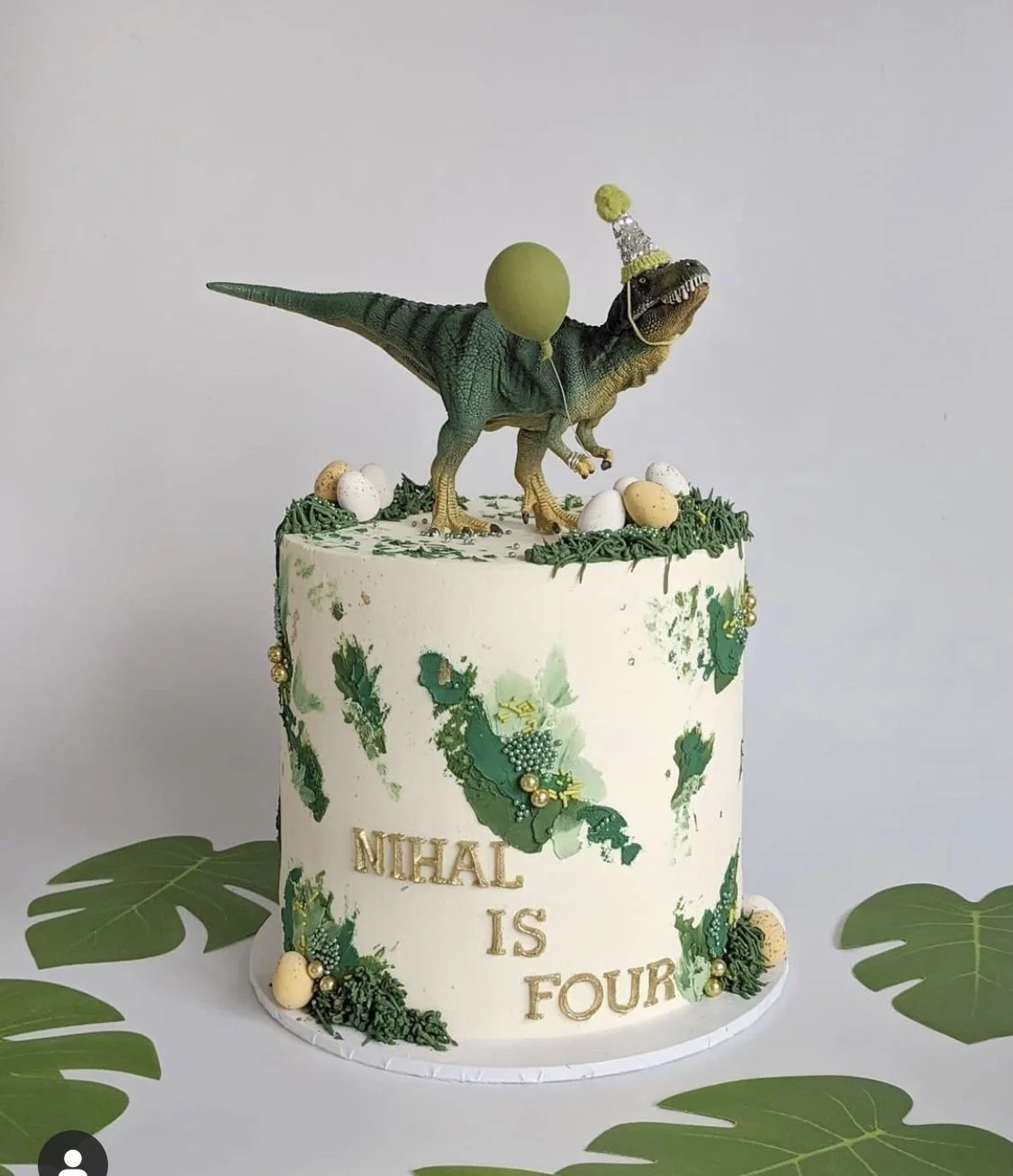 Pastel de cumpleaños con decoraciones de dinosaurios en tonos verdes y dorados, con un dinosaurio en la parte superior usando un gorro de fiesta y sosteniendo un globo verde.