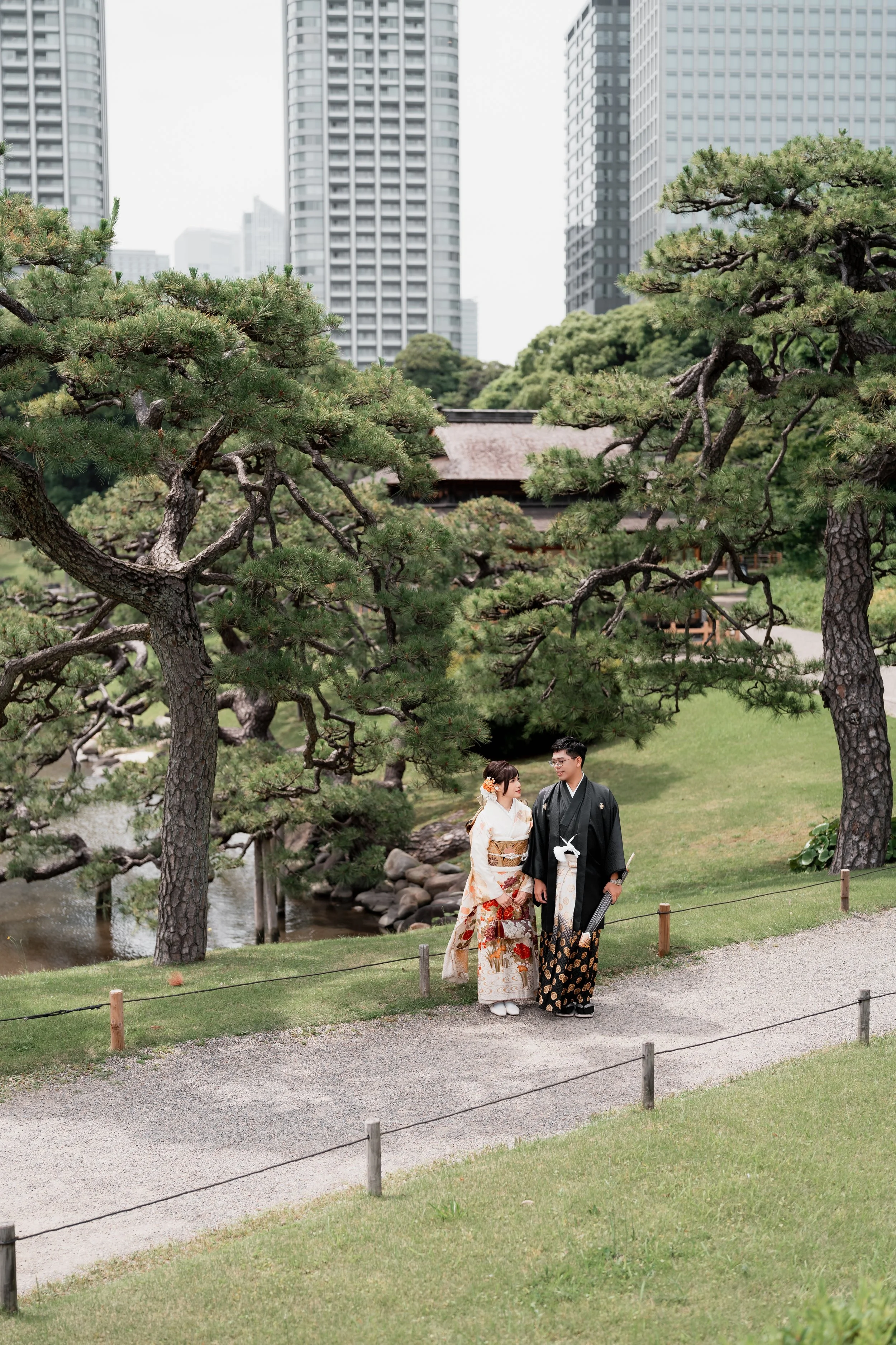 A couple dressed in traditional Japanese wedding attire standing on a path in a park with green trees and a small stream, with modern skyscrapers in the background.