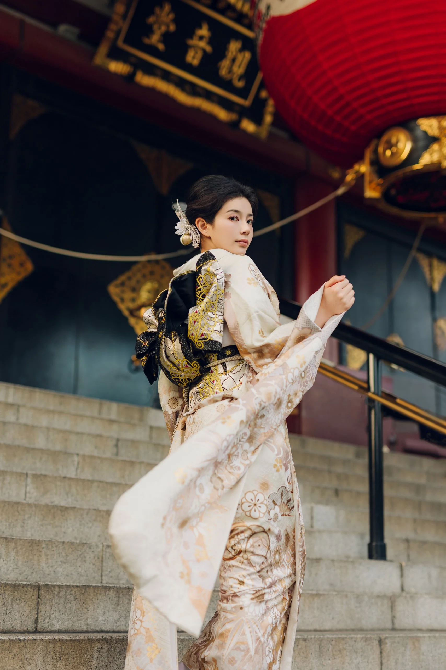 A young woman wearing a traditional Japanese kimono stands on stone steps outside a building with Asian decor, including a red lantern and gold accents.