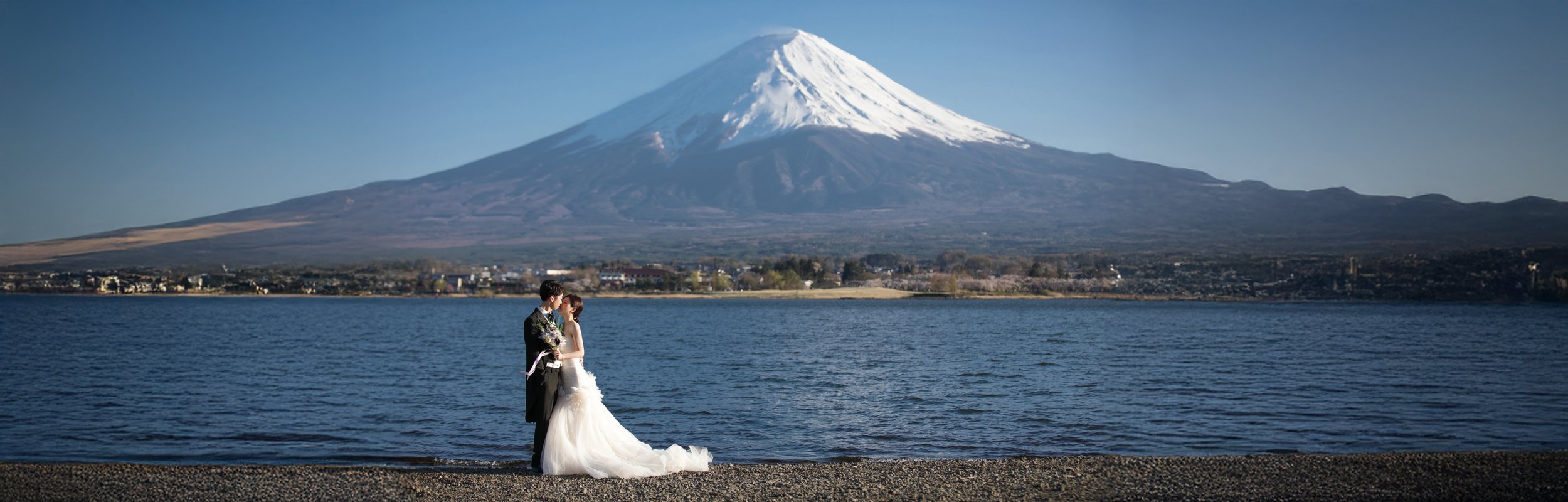 A couple in wedding attire standing by a lake, with Mount Fuji and snow-capped peak in the background.