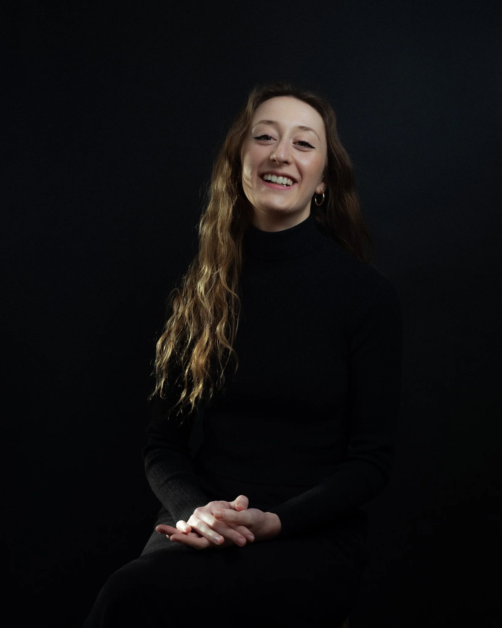 A young woman with long, wavy hair, seated, smiling, wearing a black turtleneck and hoop earrings, against a dark background.