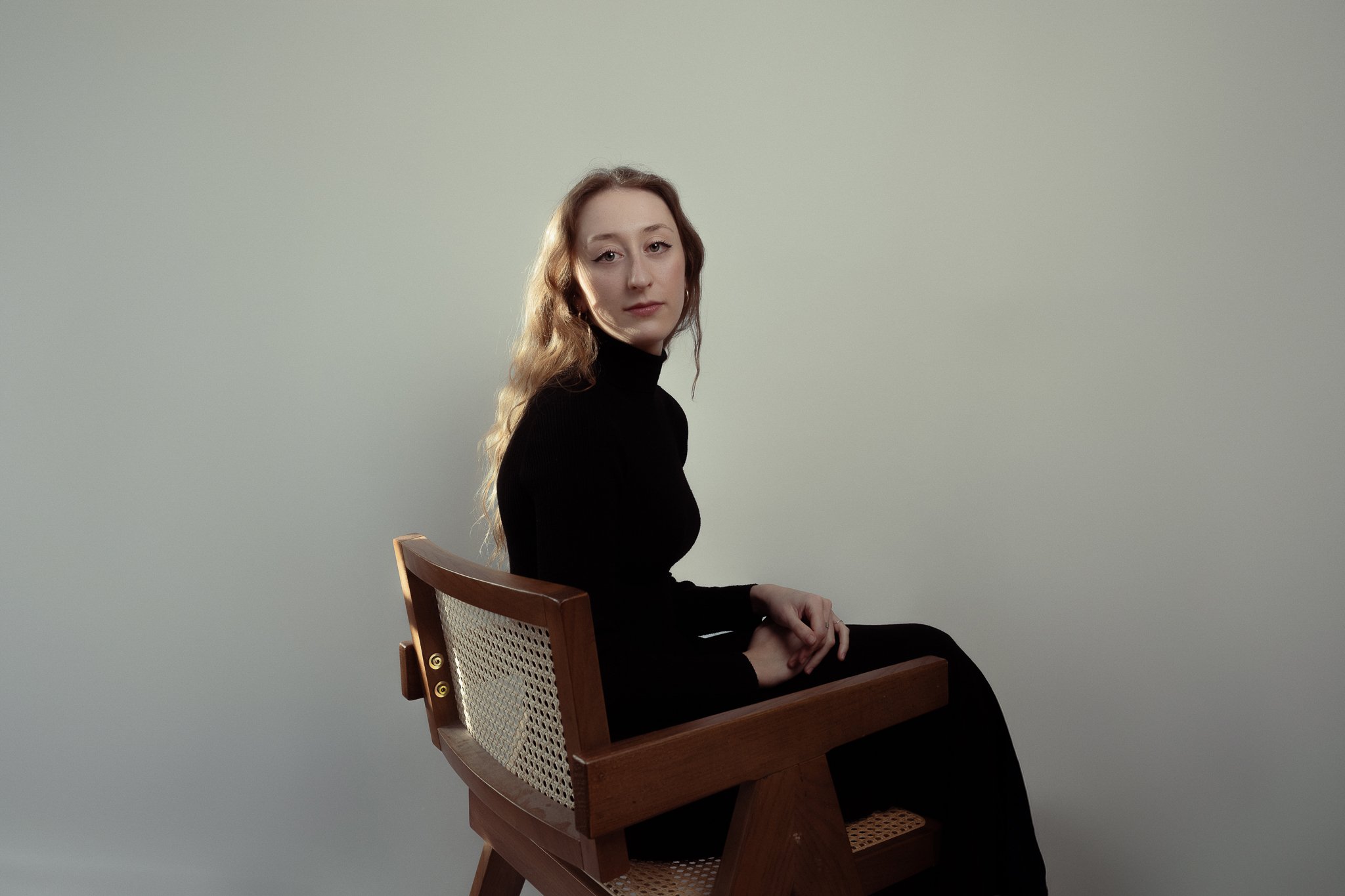 A woman with long wavy hair sitting sideways on a wooden chair, wearing a black turtleneck and black pants against a plain light-colored background.