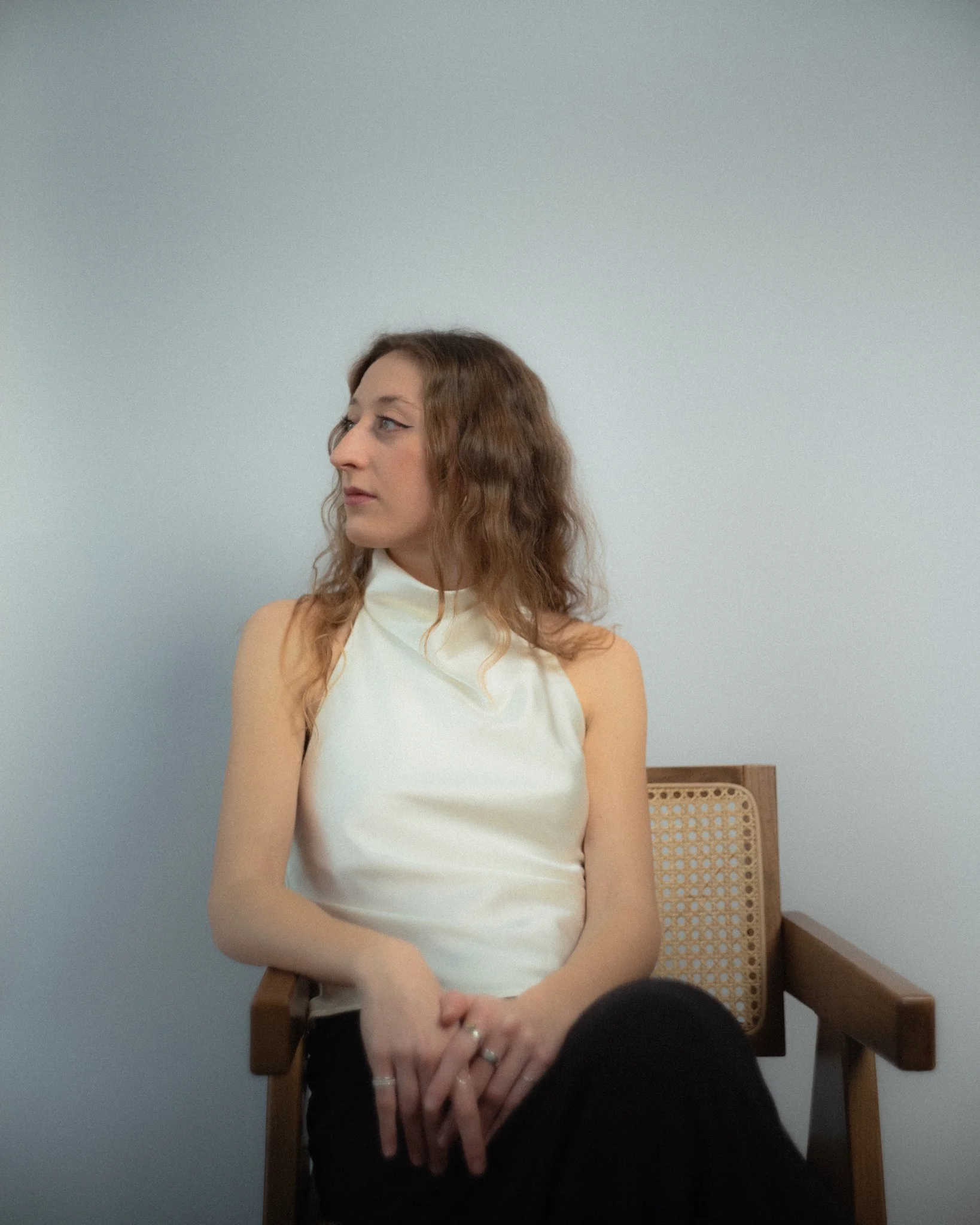 Woman with wavy brown hair wearing a sleeveless white top, sitting on a wooden chair with a cane backrest against a plain light-colored wall.