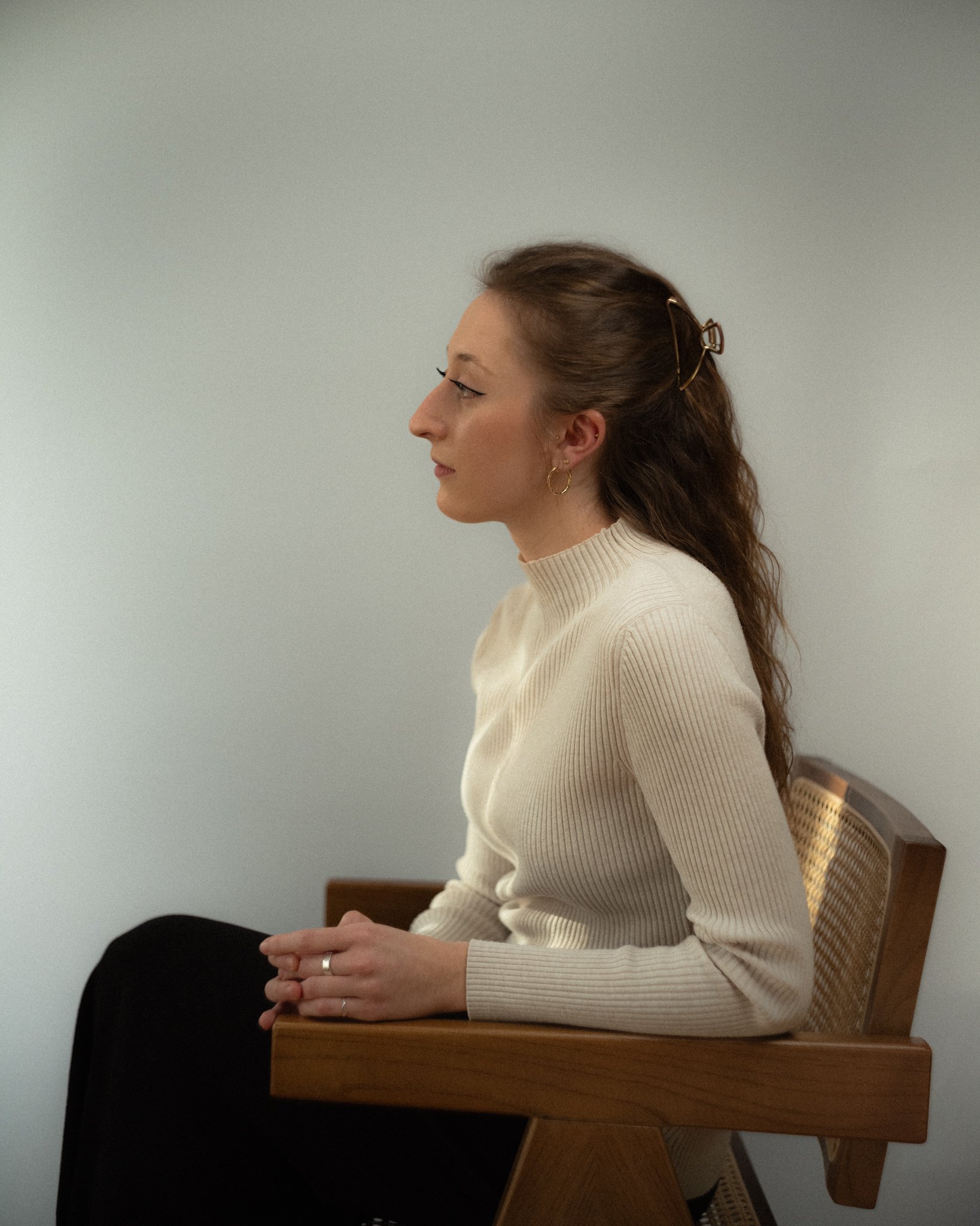 A woman with light skin, long wavy brown hair held with a hair clip, sitting sideways on a wooden chair against a plain light wall, wearing a cream-colored ribbed turtleneck sweater and gold hoop earrings.