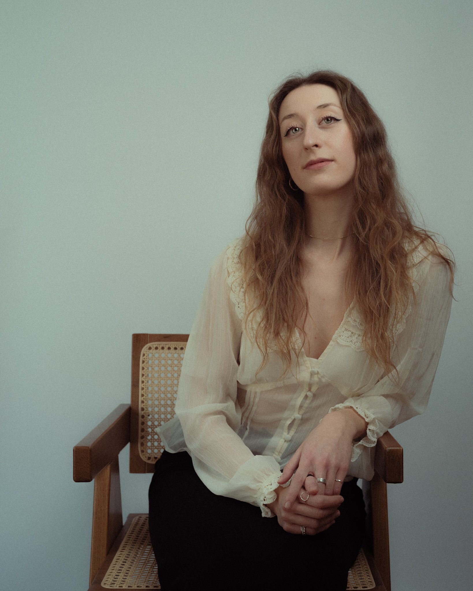 A woman with long wavy reddish-brown hair seated on a wooden chair with a cane backrest against a plain light-colored wall, wearing a cream-colored blouse with lace details.