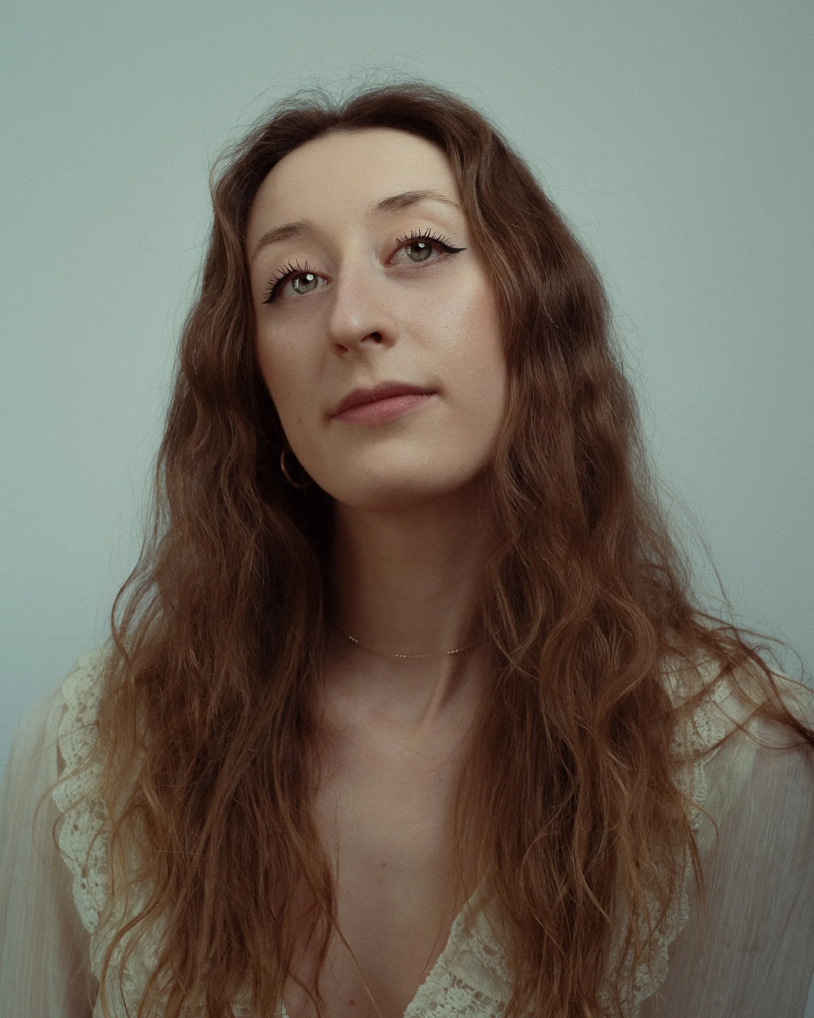 A woman with long, wavy auburn hair and light skin looking slightly upward, wearing a light-colored top with lace details and a delicate gold necklace, against a plain light gray background.