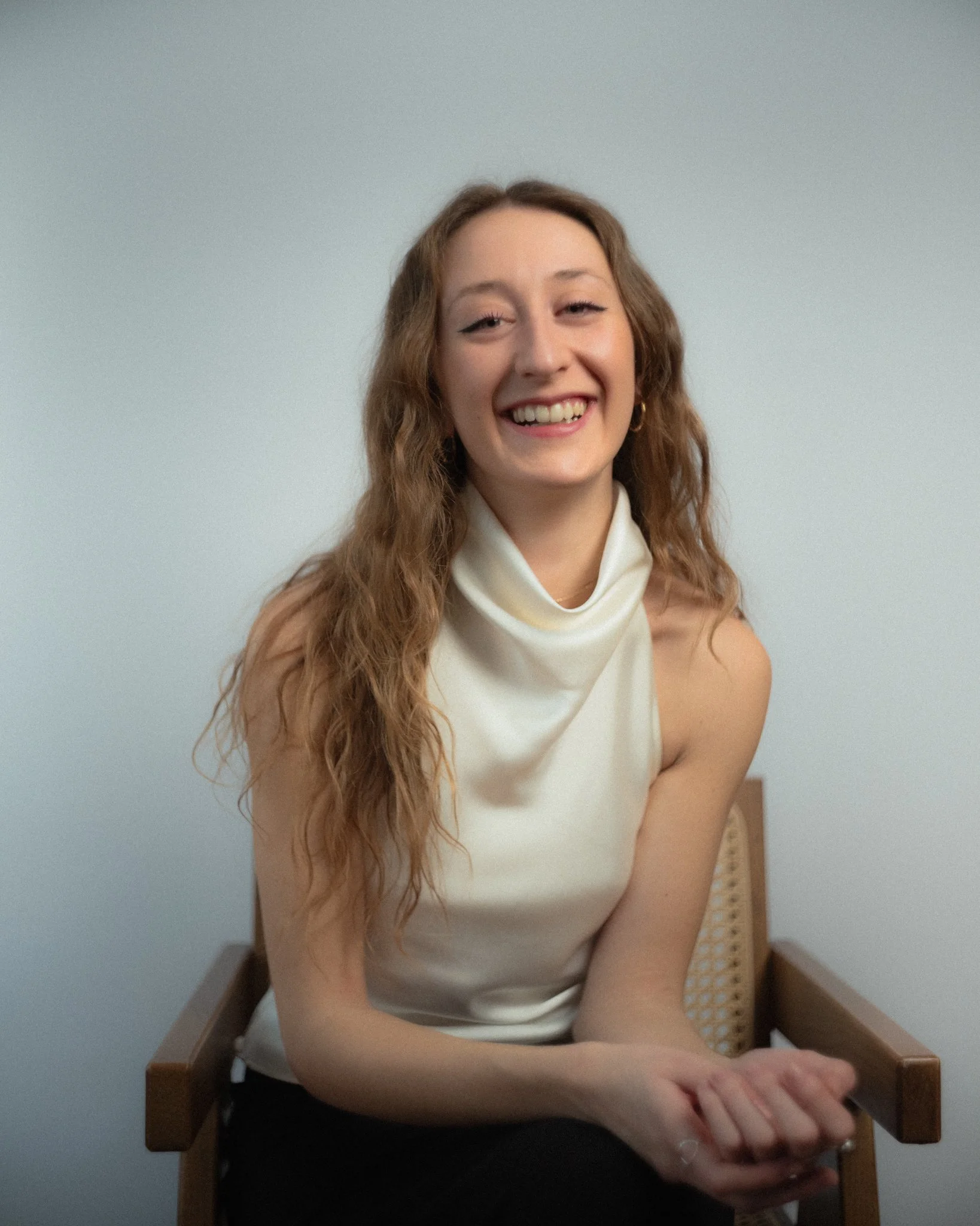 A woman with long, wavy red hair, smiling, wearing a white sleeveless top and sitting on a wooden chair.
