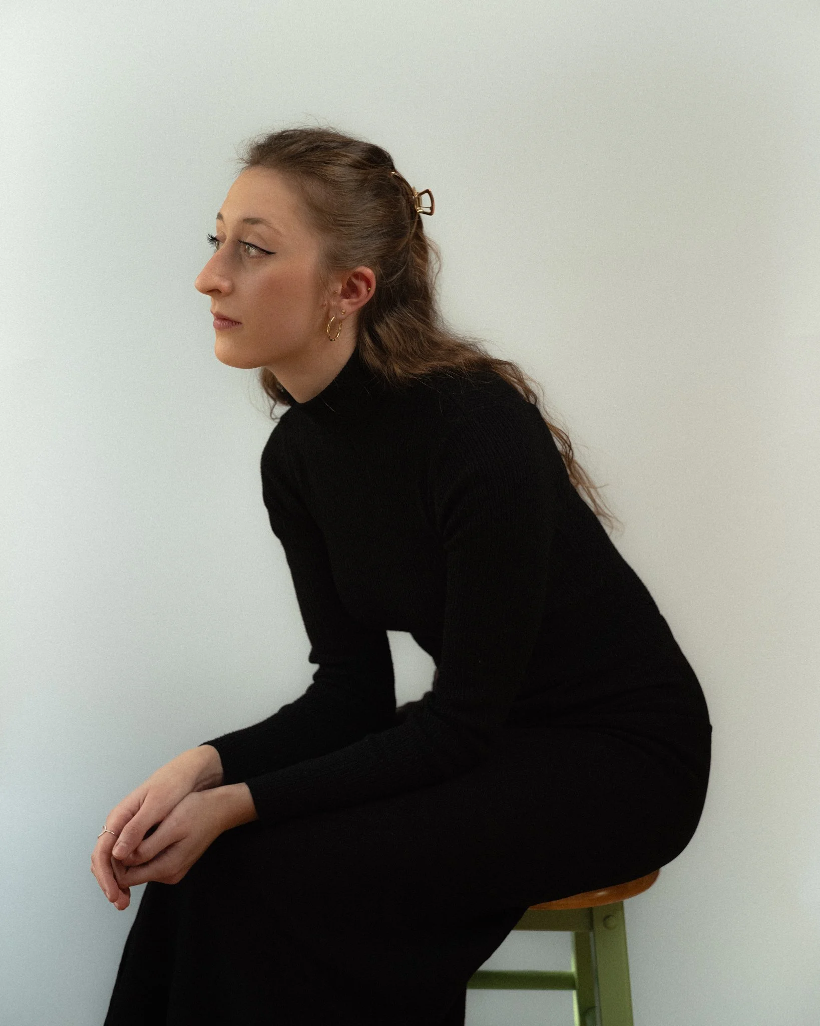 A woman in black sits on a green chair against a plain white wall, with her hands clasped and looking to the left.