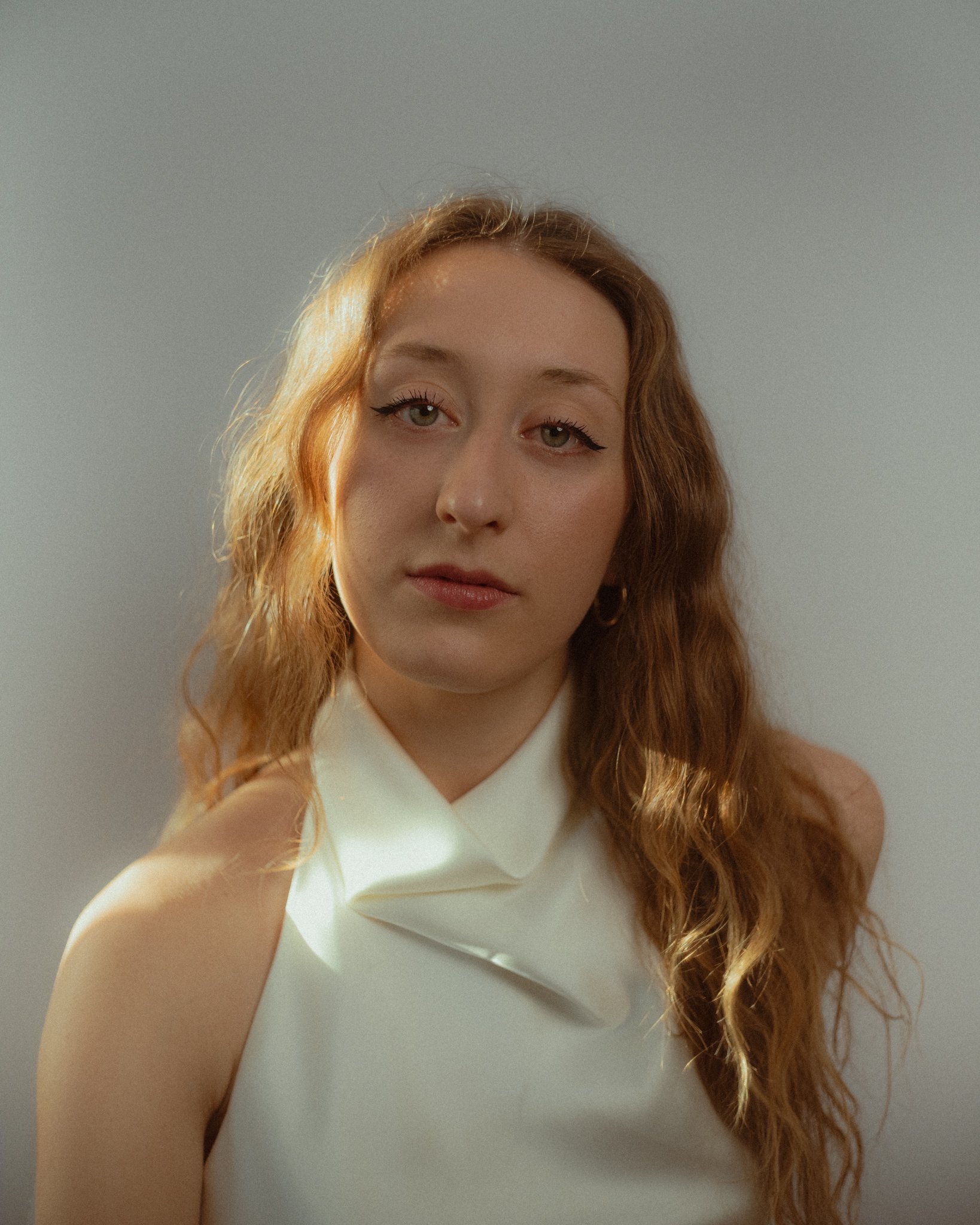 A young woman with wavy red hair wearing a sleeveless white collared top, looking at the camera with a neutral expression, standing against a plain background.