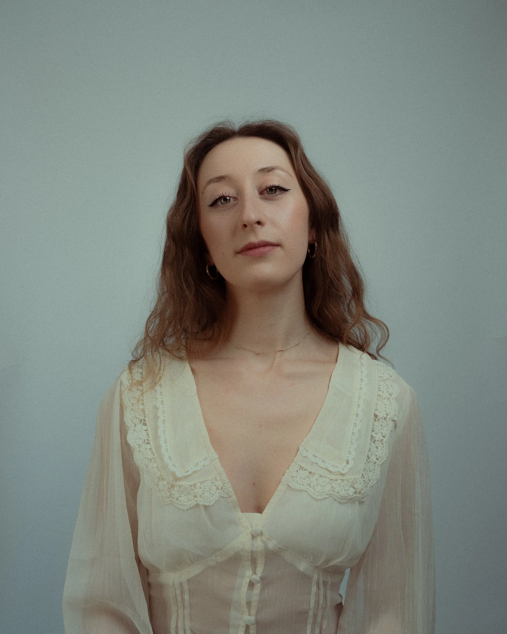 A woman with long wavy brown hair and fair skin looking directly at the camera, wearing a cream-colored blouse with lace details and a V-neck, standing against a plain light blue background.