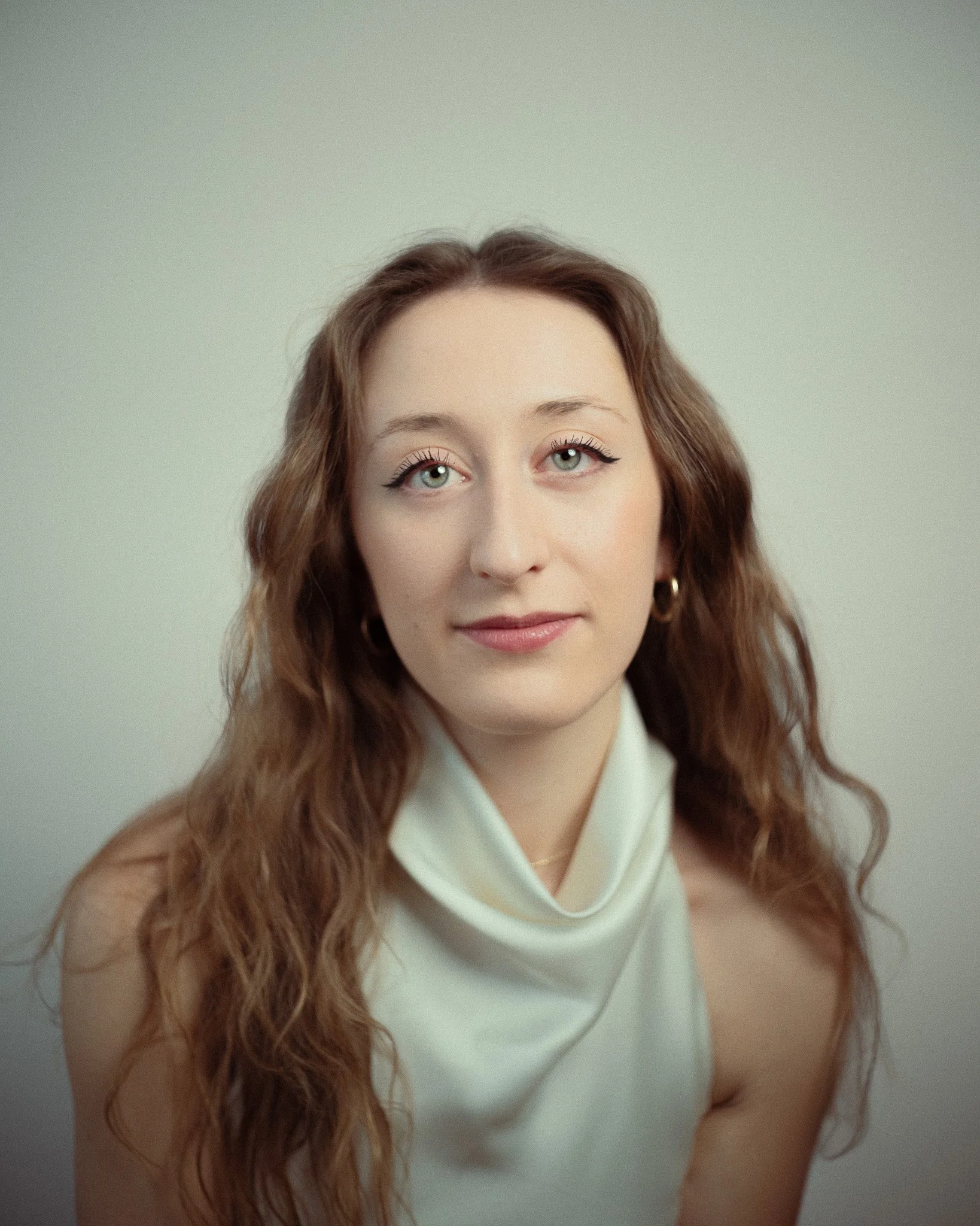 Portrait of a young woman with wavy brown hair, light skin, and blue eyes wearing a white high-neck top and gold hoop earrings, against a plain background.