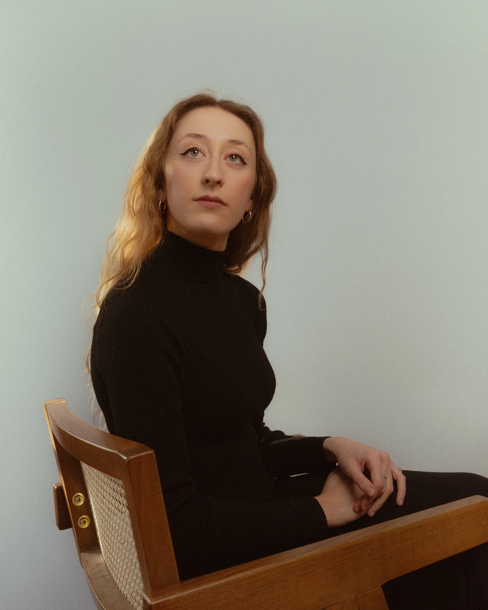 A woman with wavy reddish hair and hoop earrings sitting on a wooden chair against a plain light-colored background, wearing a black turtleneck sweater.