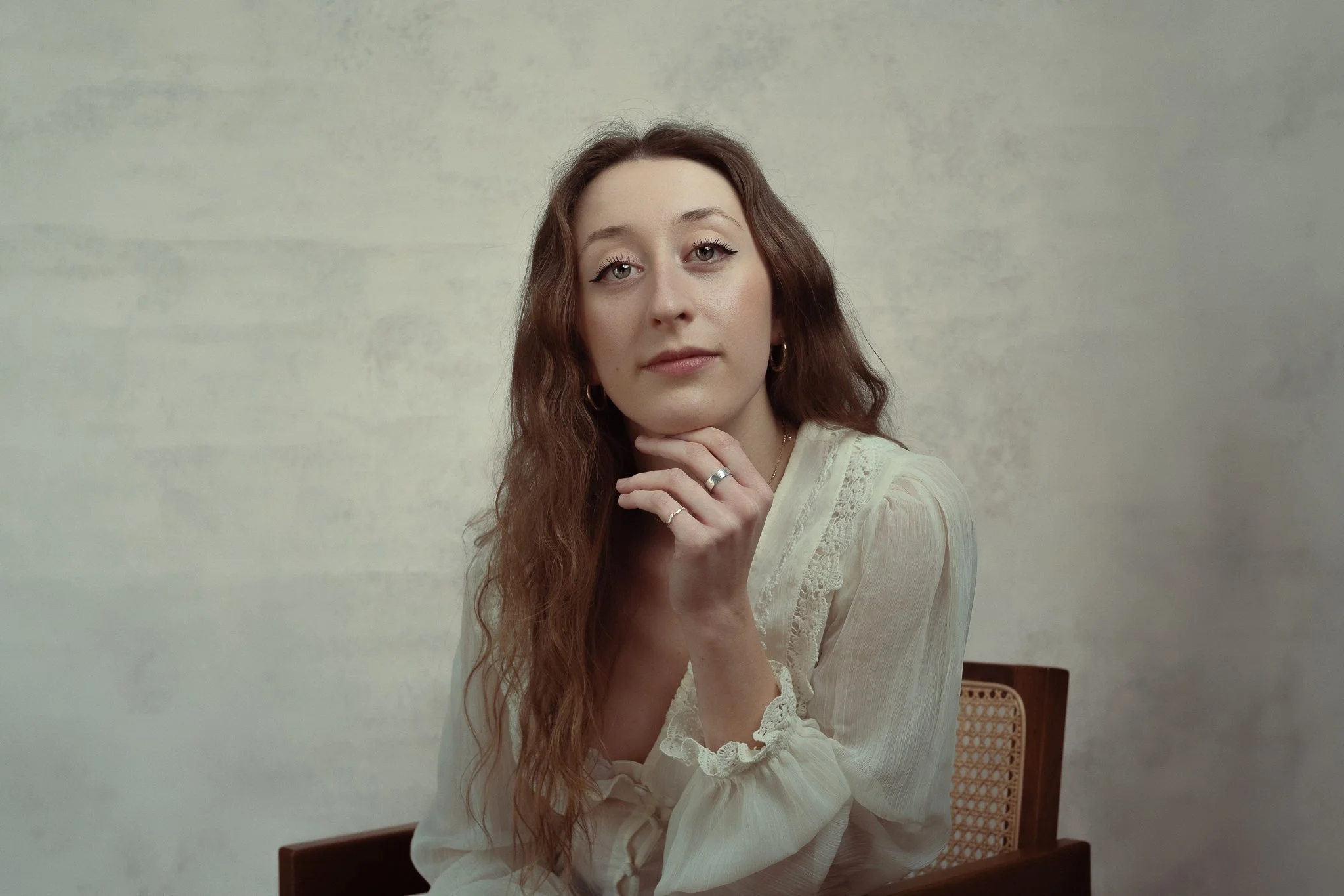 A young woman with long, wavy brown hair, wearing a cream-colored blouse with lace details, is sitting on a wooden chair with a woven backrest. She has her left hand near her chin, showing rings on her fingers, and is looking directly at the camera w