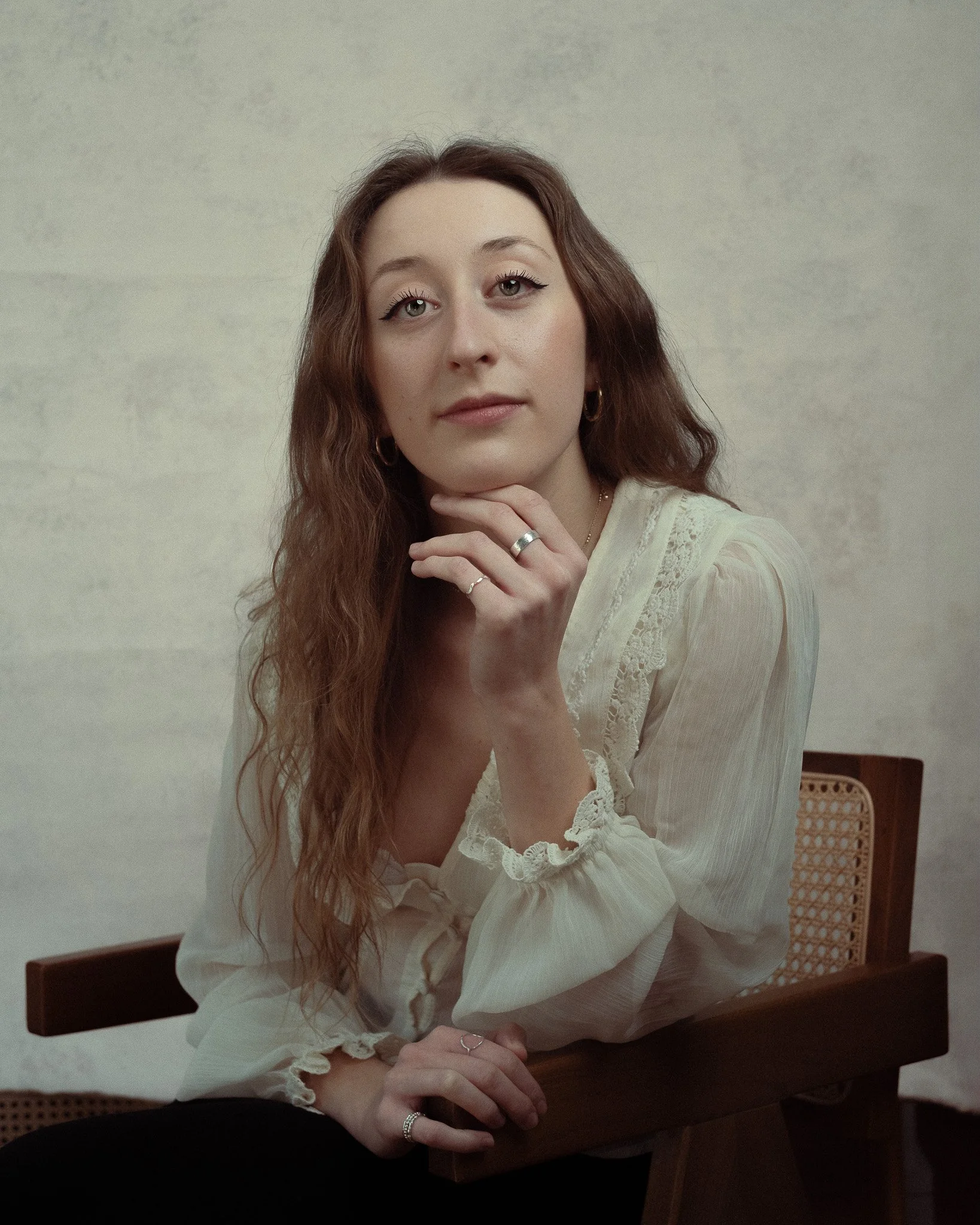 A woman with long, wavy auburn hair, wearing a cream-colored blouse with lace details, seated on a chair with a woven backrest against a plain light-colored background.