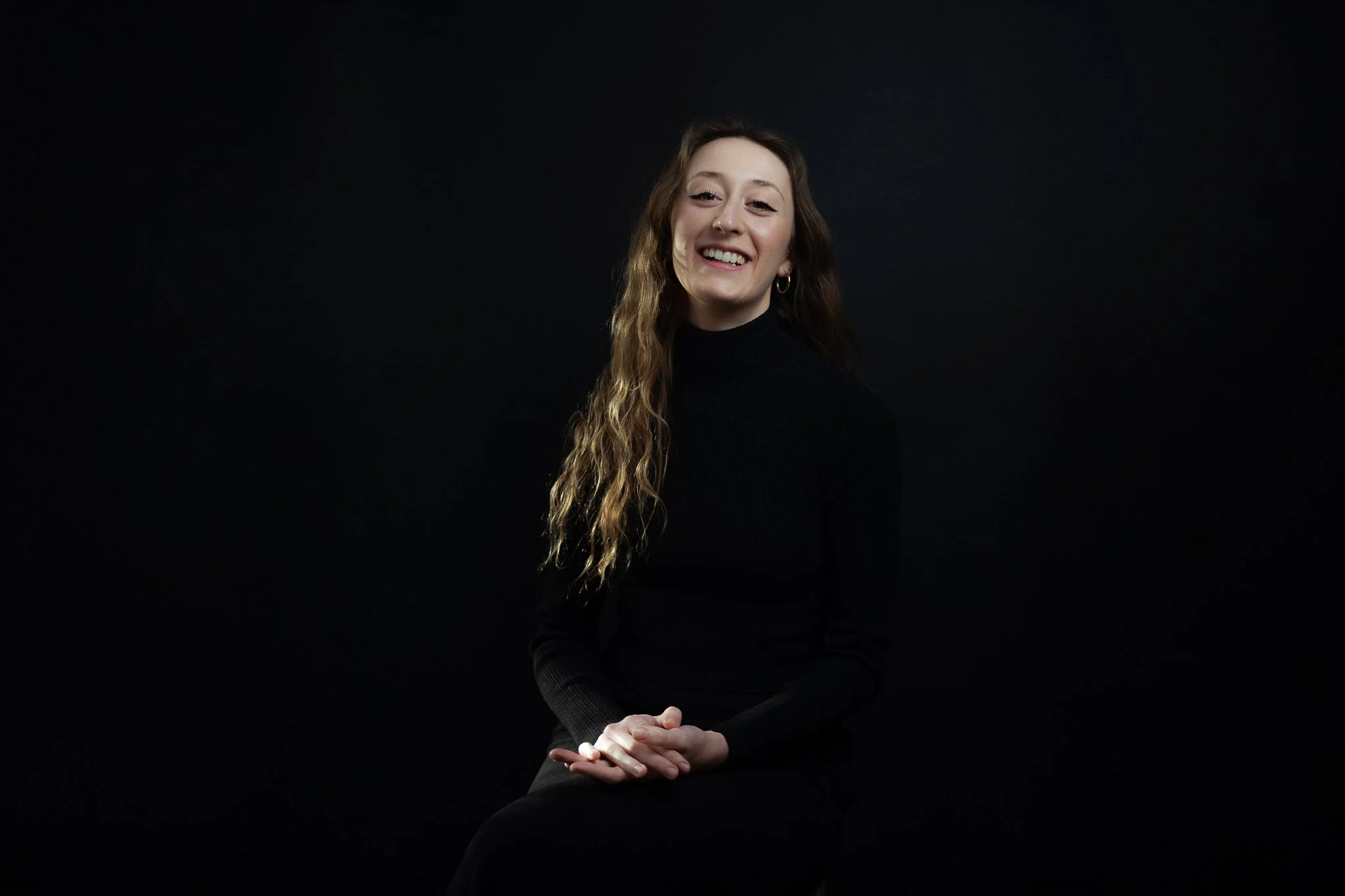 A woman with long wavy hair wearing a black turtleneck and earrings, smiling and sitting against a dark background.