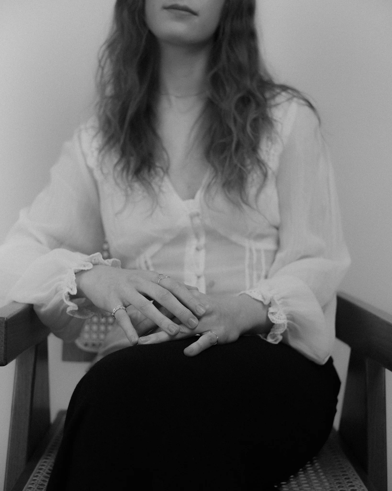 A woman sitting on a chair with her hands resting on her lap, wearing a light-colored blouse and multiple rings on her fingers.