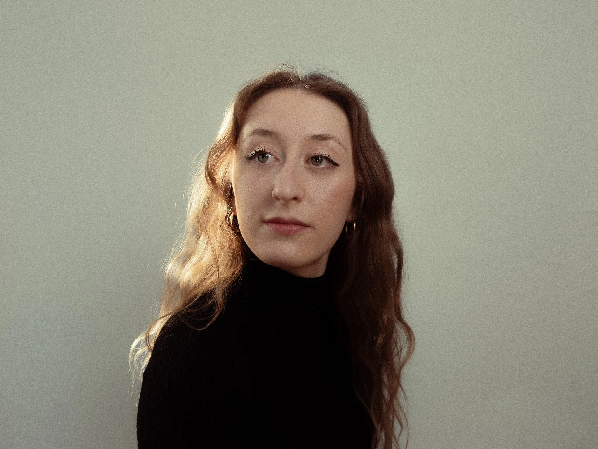 A woman with long, wavy, reddish-brown hair wearing a black turtleneck, looking to her left against a light gray background.