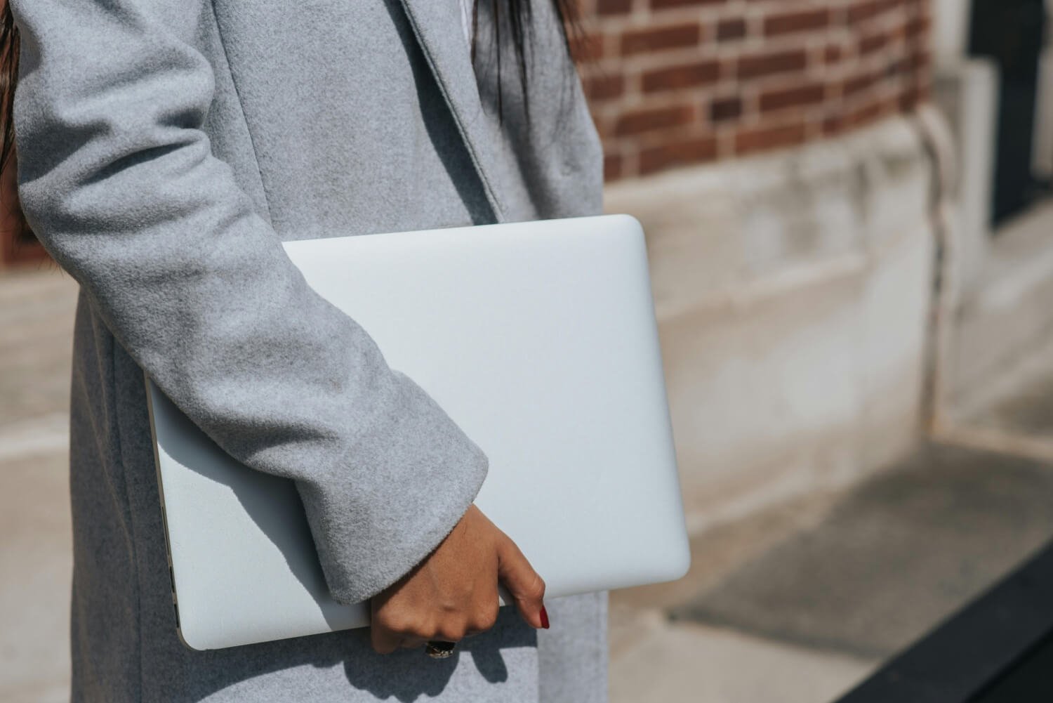 Person wearing a gray coat holding a closed laptop in front of a brick wall.