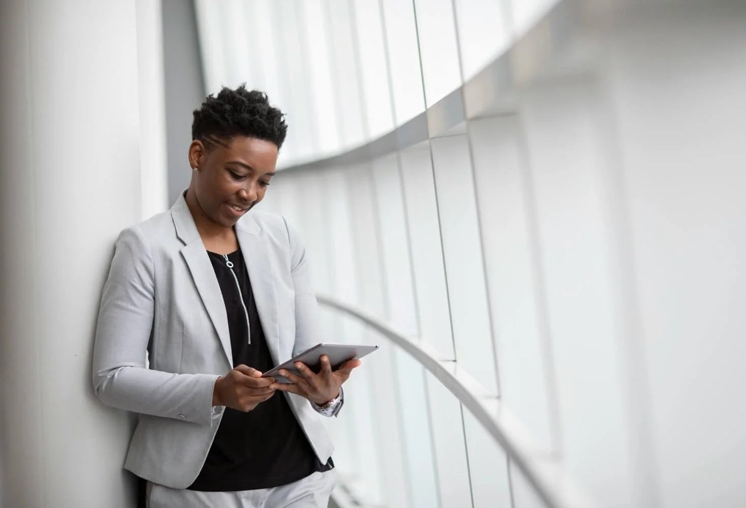 A woman with short curly hair wearing a light gray blazer and black top, leaning against a wall while looking at a tablet in her hands.