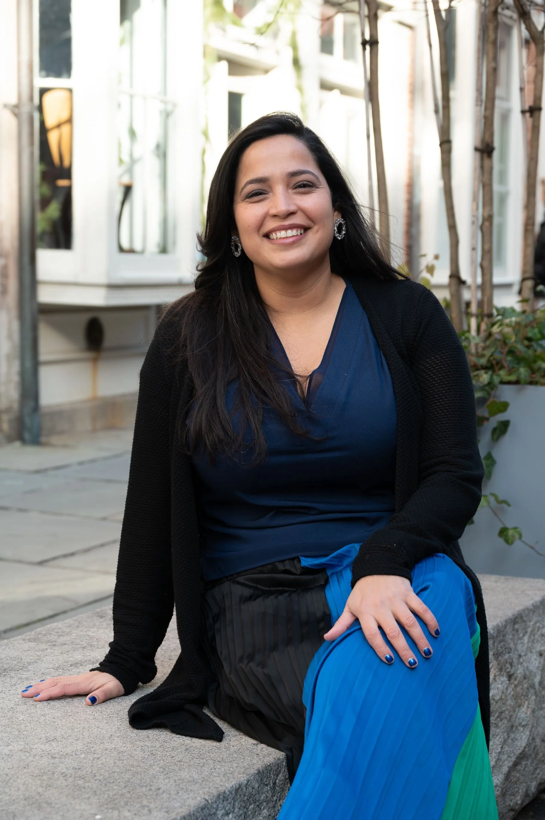 Nadia Hernandez sitting outdoors on a stone bench, smiling, wearing a blue top, black cardigan, and a colorful blue and green skirt.