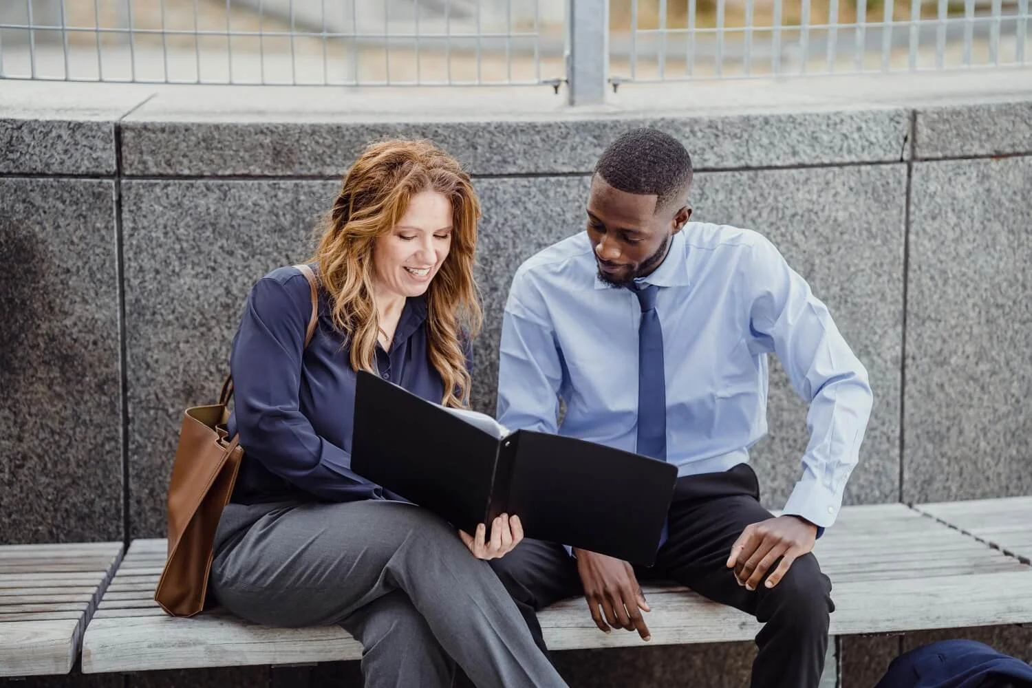 A woman and a man sitting on a bench outdoors, looking at a black photo album together and smiling.