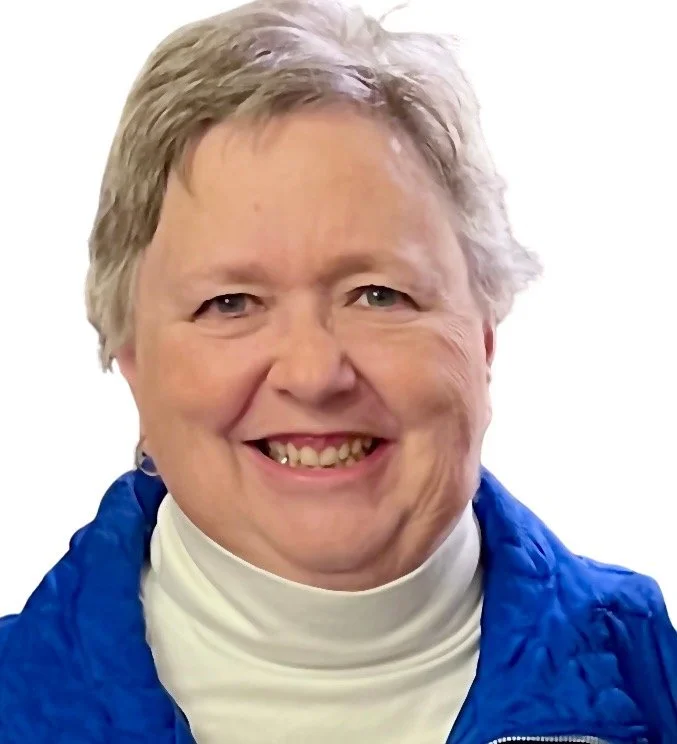 A smiling woman with natural curly hair, wearing glasses, hoop earrings, layered necklaces, and a striped shirt.