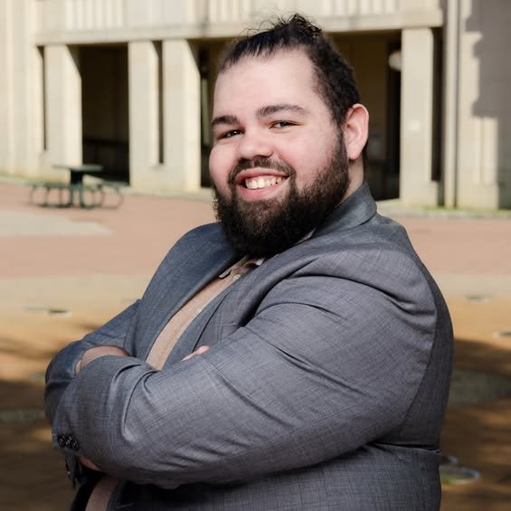 A man with glasses, a beard, and curly dark hair, looking to the side with a slight smile, wearing a dark green sweater, sitting indoors against a neutral background with a plant in the background.