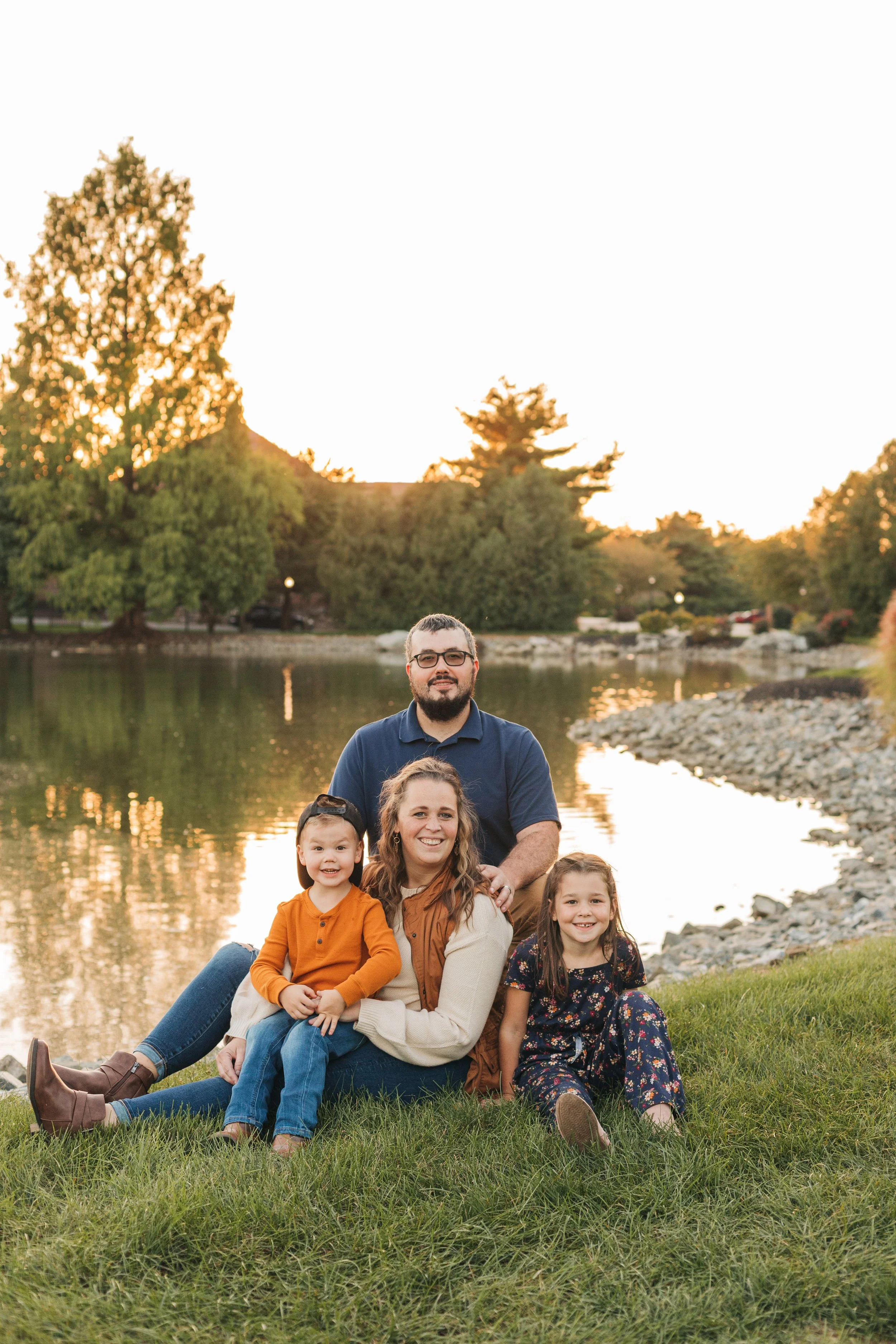 A family of four sitting on grass by a lake during sunset, smiling at the camera.