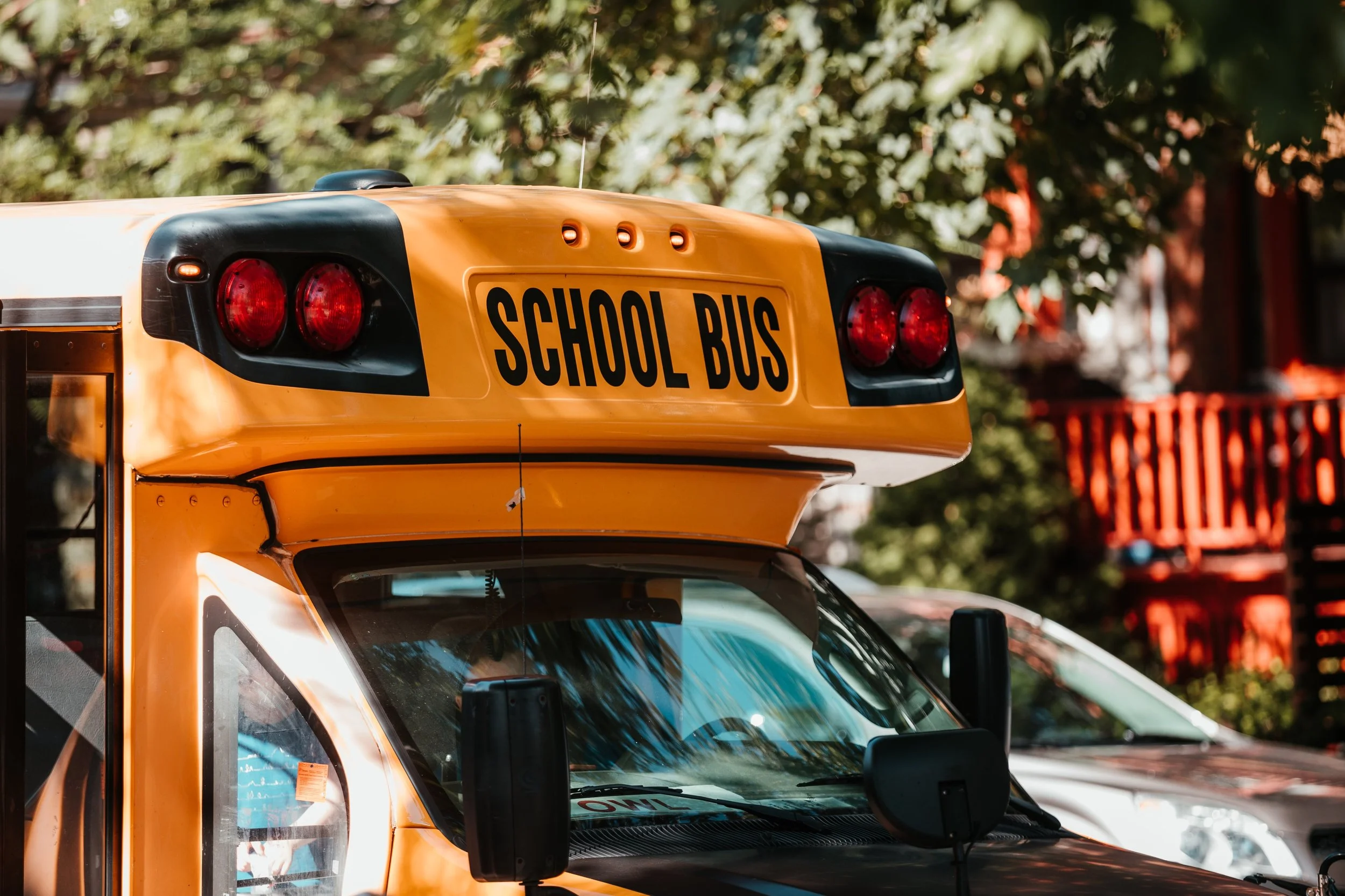 Close-up view of the front of a yellow school bus with a sign that says 'SCHOOL BUS' on the top front, parked outdoors with trees and a red building in the background.