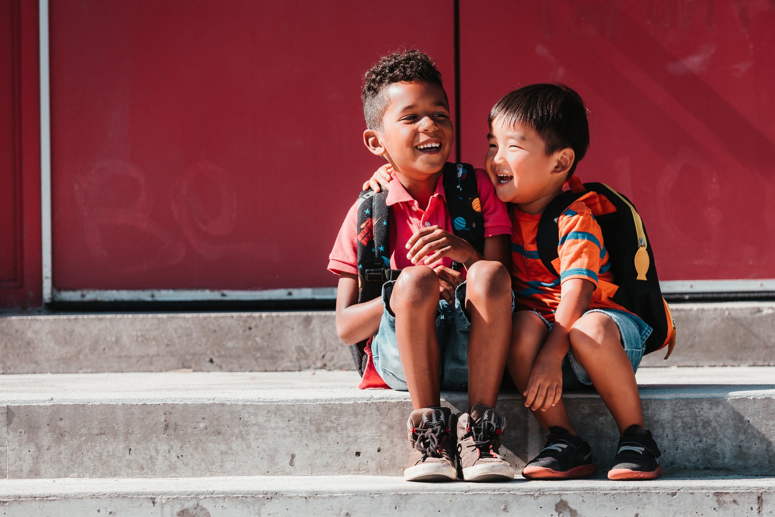 Two young boys sitting on steps, laughing and smiling, wearing backpacks, in front of a red wall.