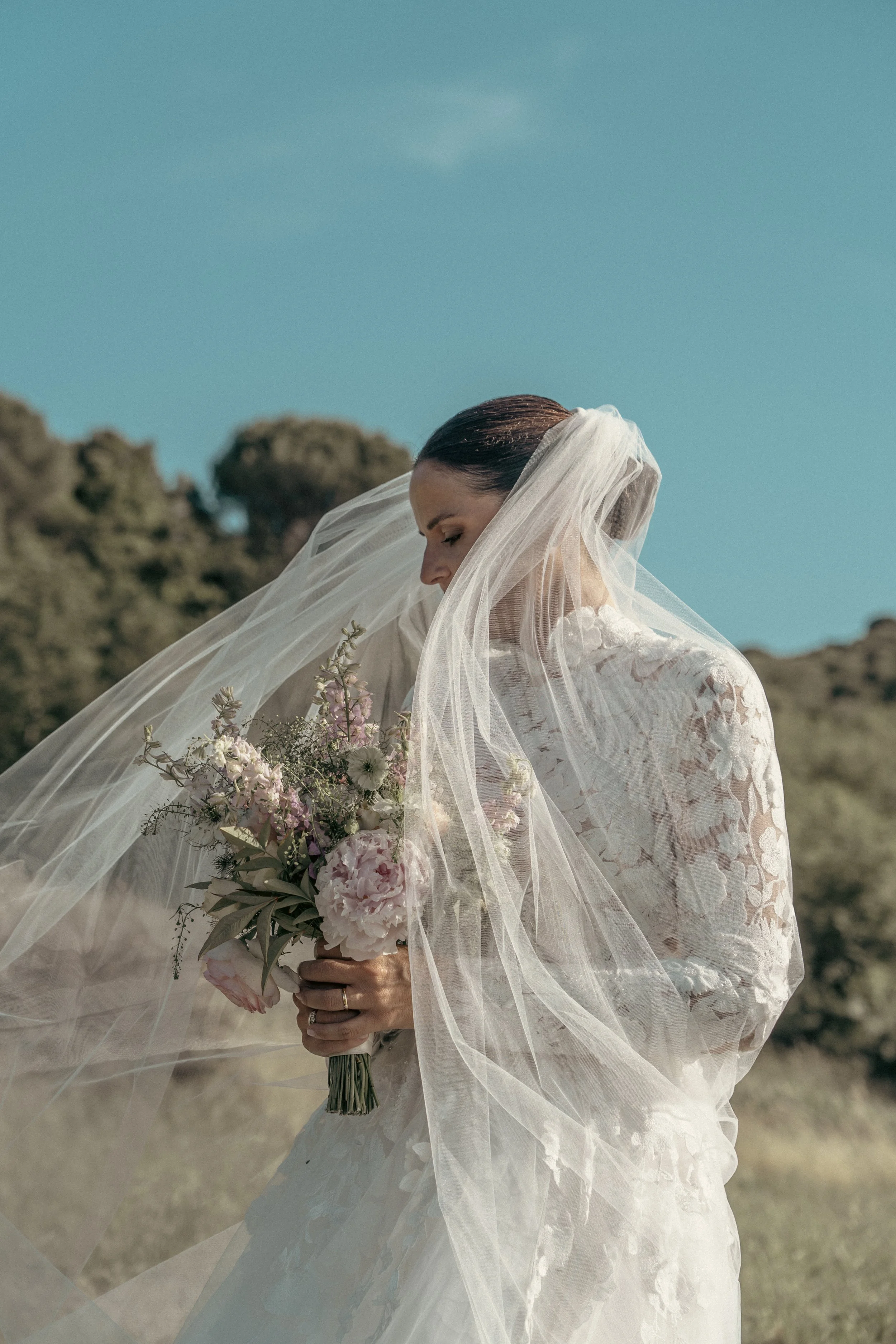 A bride in a white lace wedding dress holding a bouquet of pink and white flowers, with a veil over her head, outdoors on a sunny day.