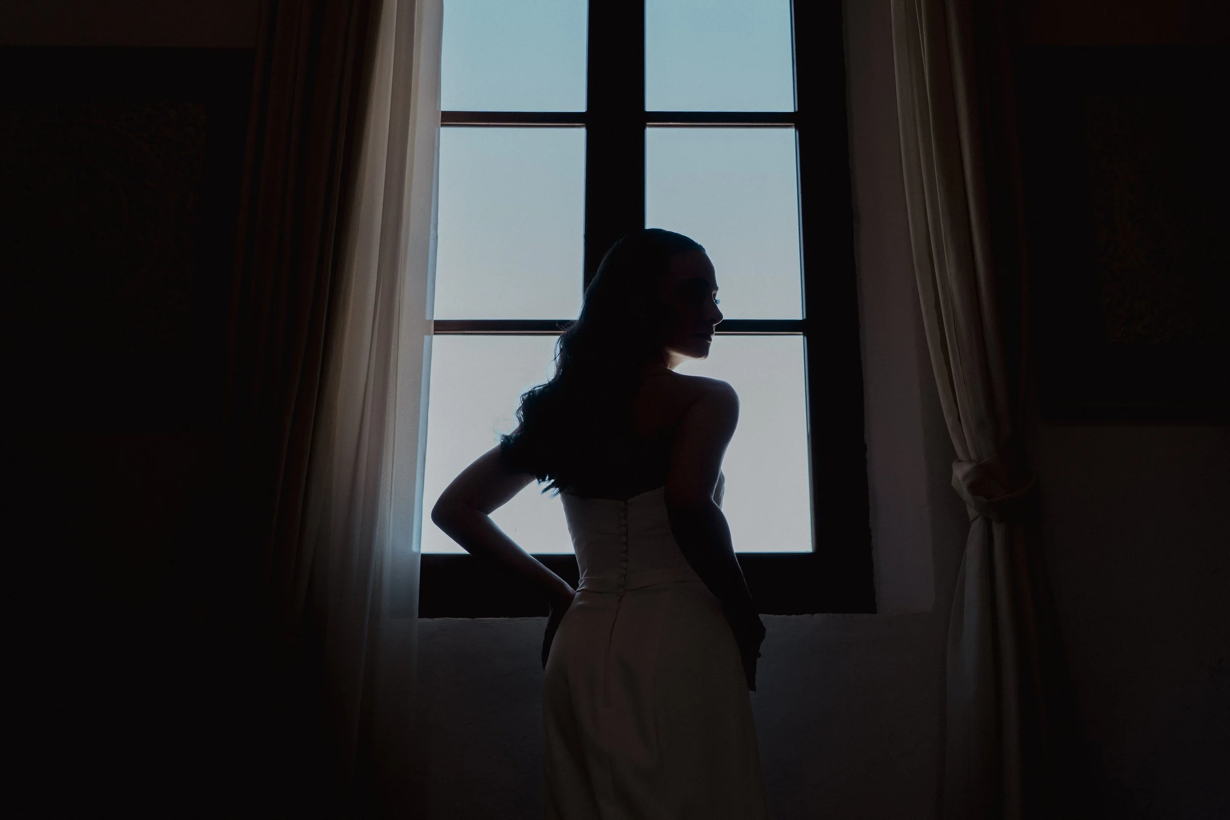 Silhouette of a woman in a strapless dress standing by a window with curtains, facing sideways.