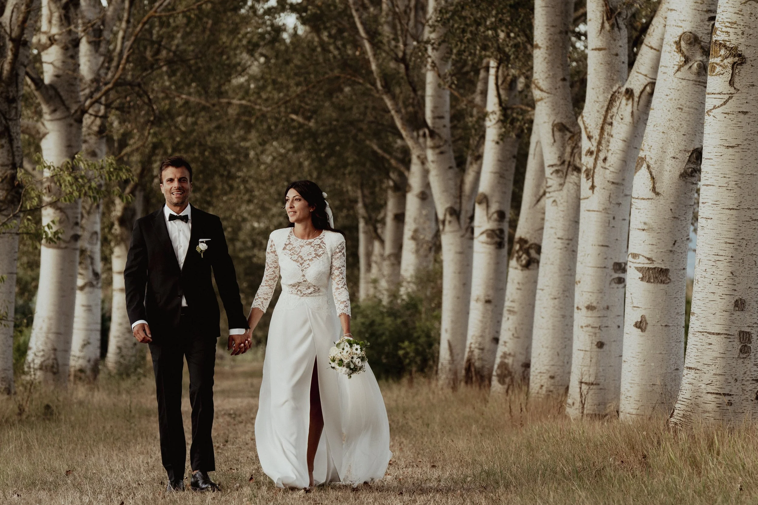 A newlywed couple holding hands and walking through a grove of white-barked trees, with the bride in a white lace wedding dress and the groom in a black tuxedo, during what appears to be a wedding photo shoot.
