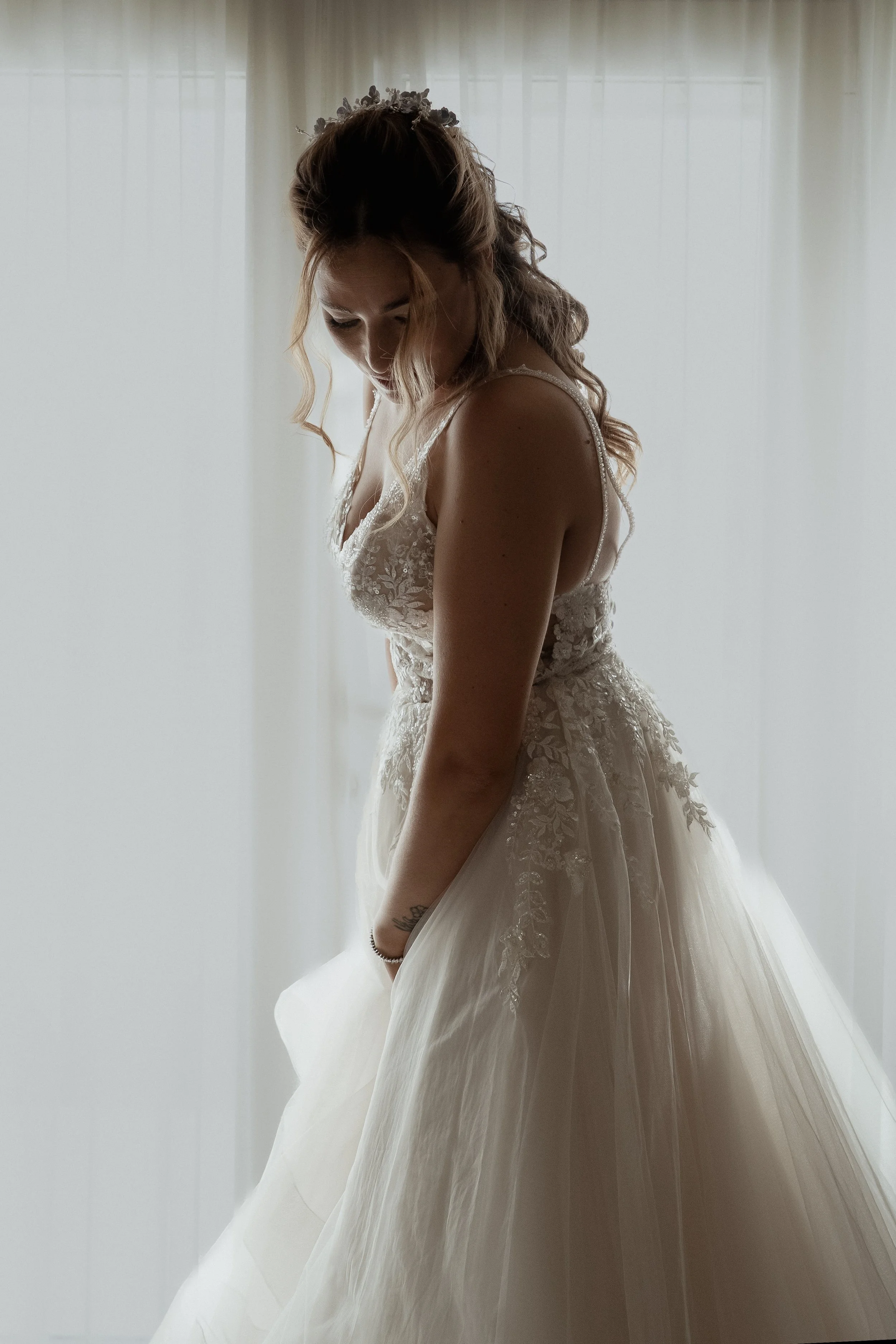 A woman in a wedding dress standing near a window, looking down, with soft natural light illuminating her.