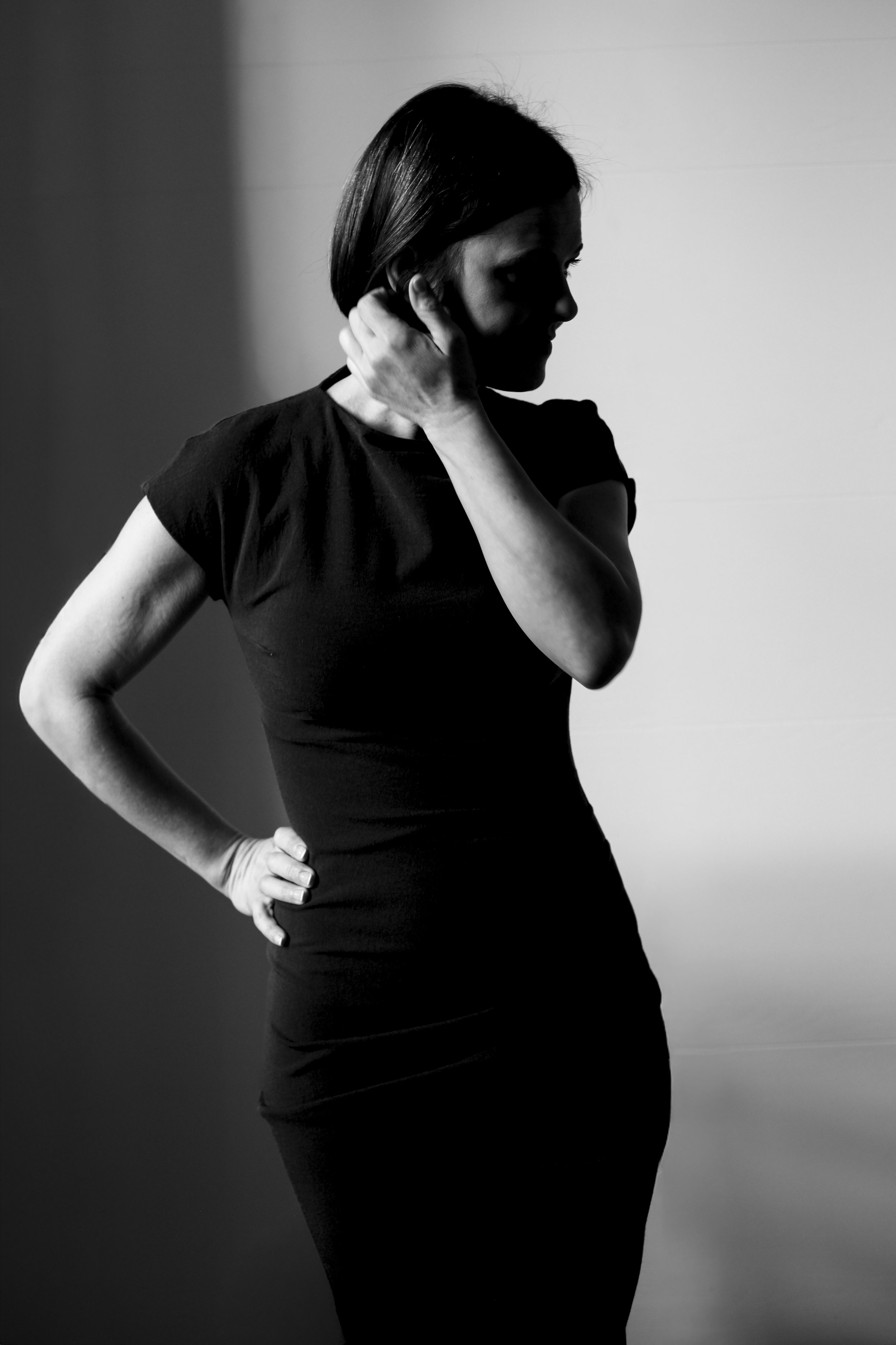 Black and white photo of a woman in a fitted black dress with her hand on her hip, standing in a shadowed room.