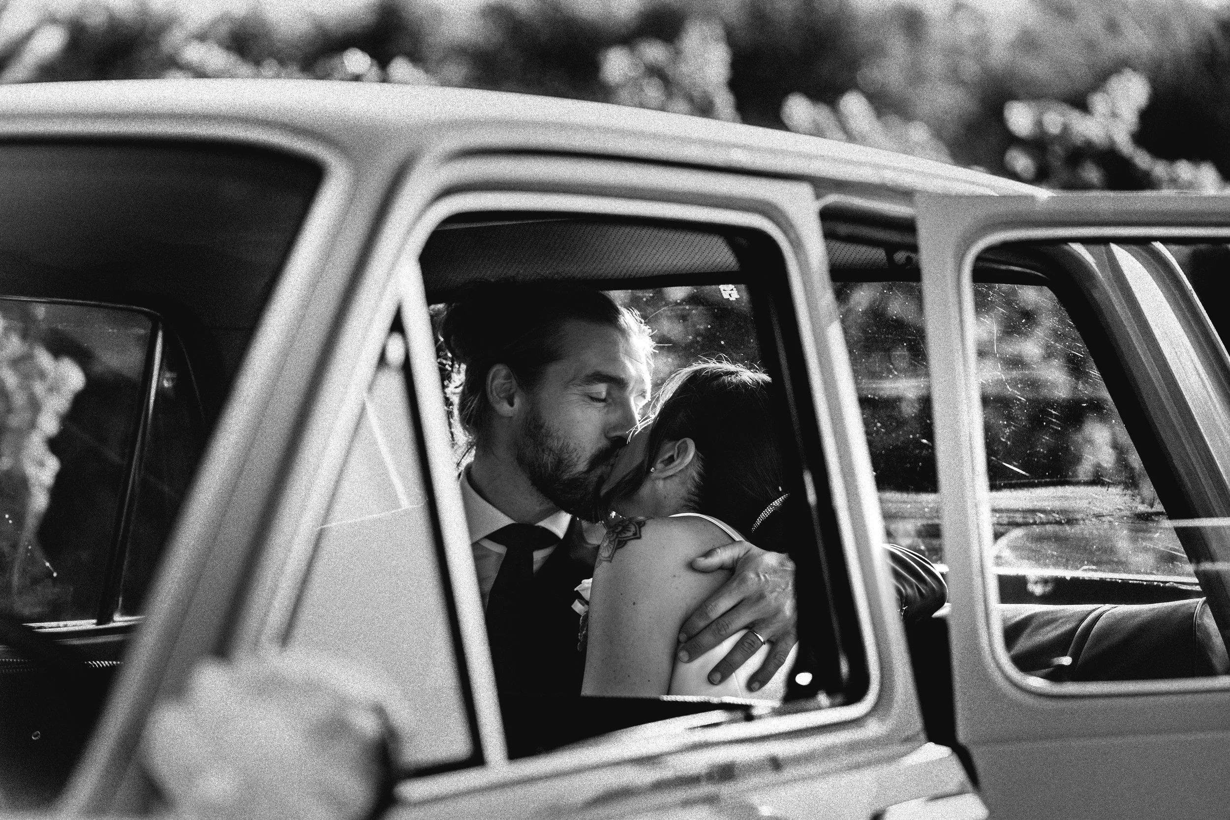 A black and white photo of a couple kissing inside a vintage car, with the man embracing the woman and their eyes closed, sunlight in the background.