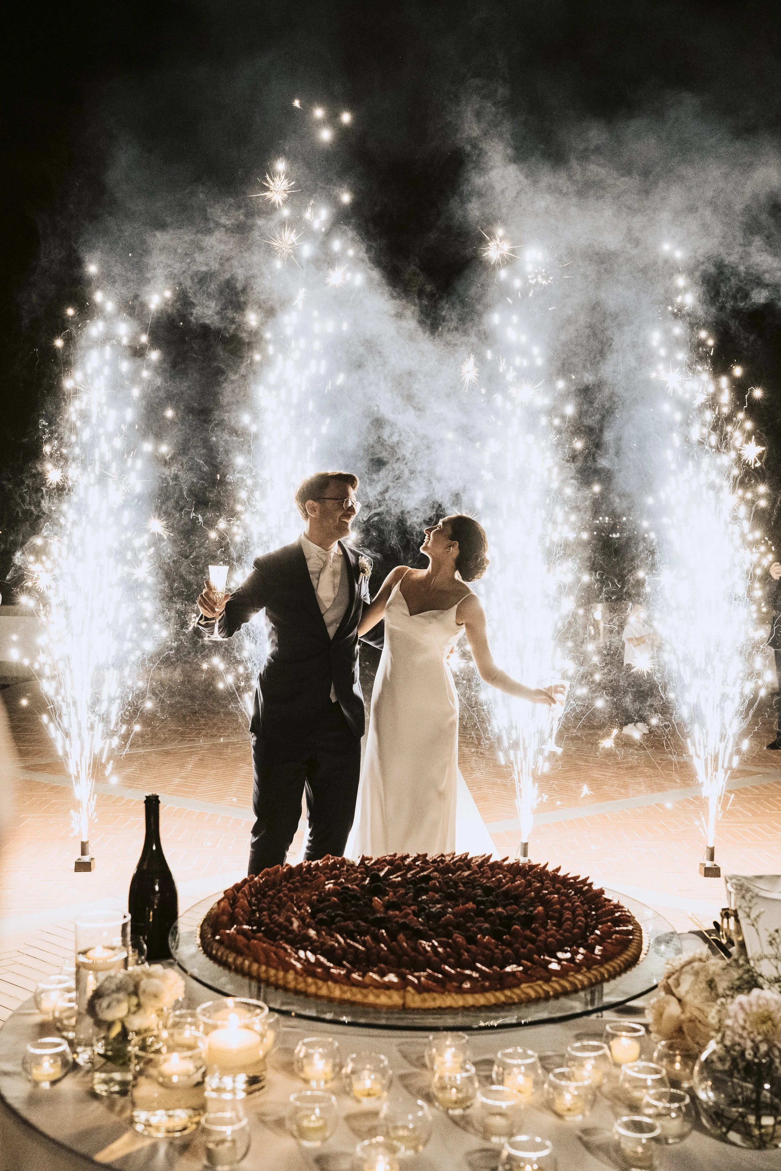 A wedding celebration with a newlywed couple dancing in front of fireworks and sparklers, with a large cake and candles on a table in the foreground.