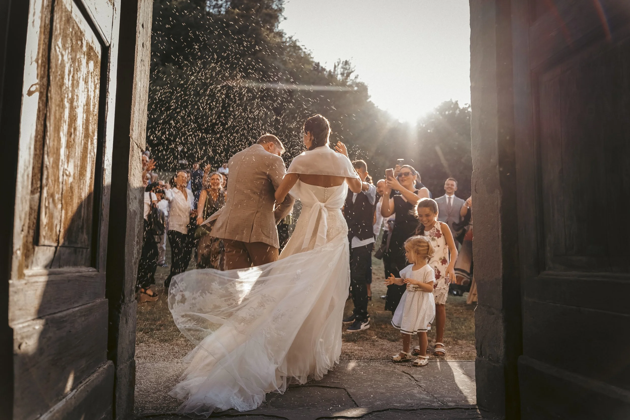 A bride and groom exit through a rustic wooden gate, surrounded by friends and family celebrating their wedding outdoors at sunset, with rice or confetti being thrown.