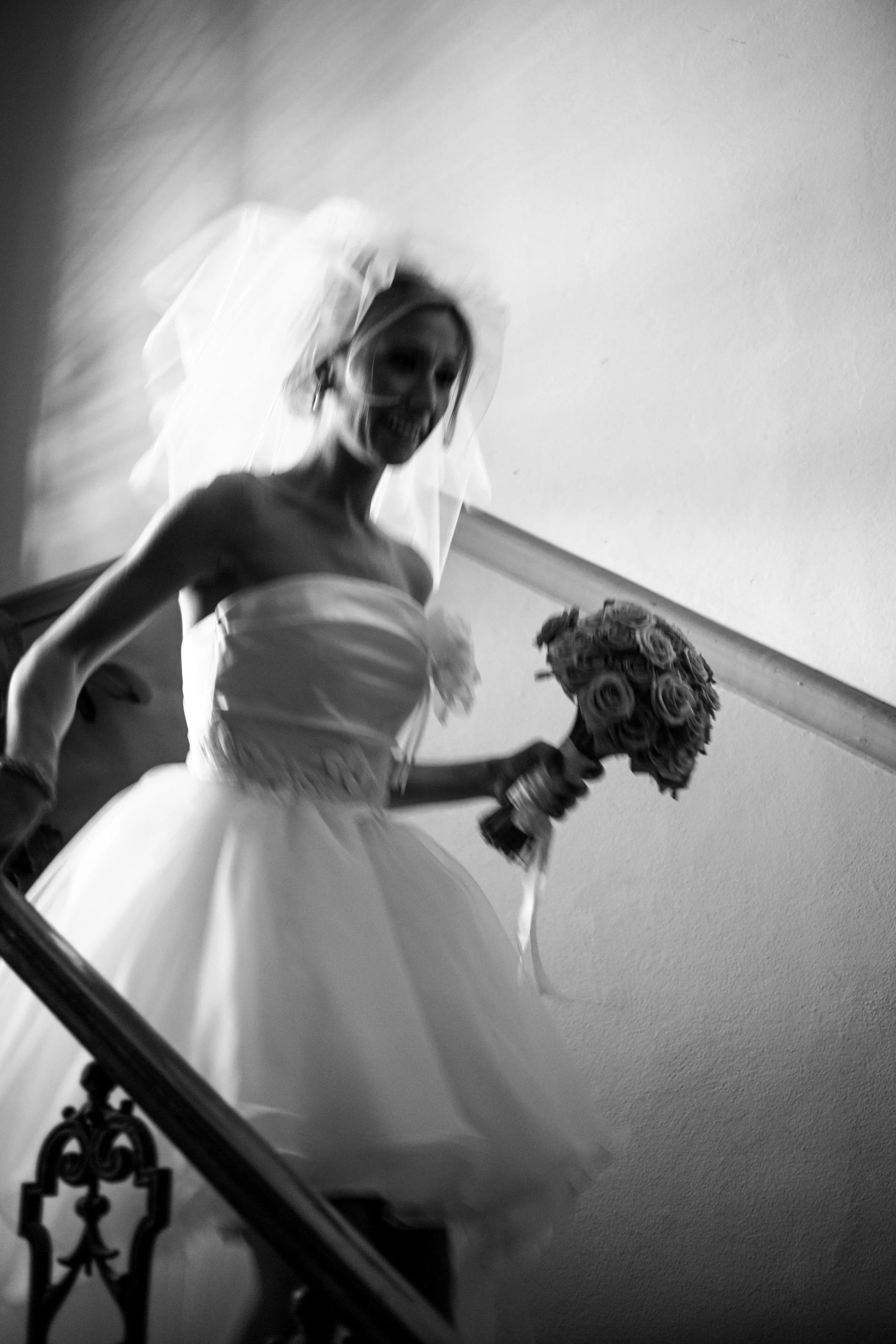 Black and white photo of a smiling bride in a strapless wedding dress, holding a bouquet of roses, ascending a staircase.