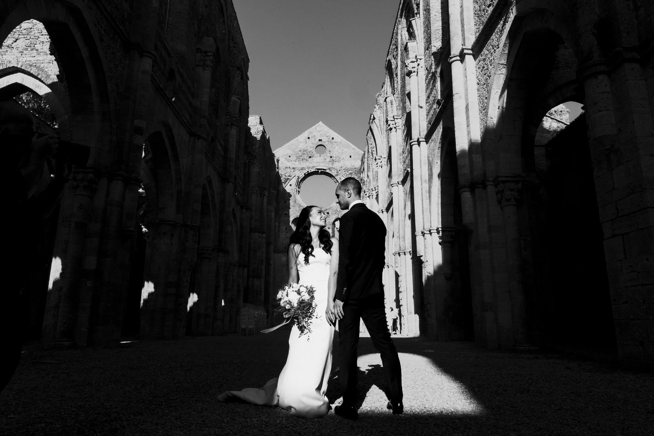 Black and white photo of a bride and groom standing close in a historic ruin with large arches and stone walls, holding hands and smiling.