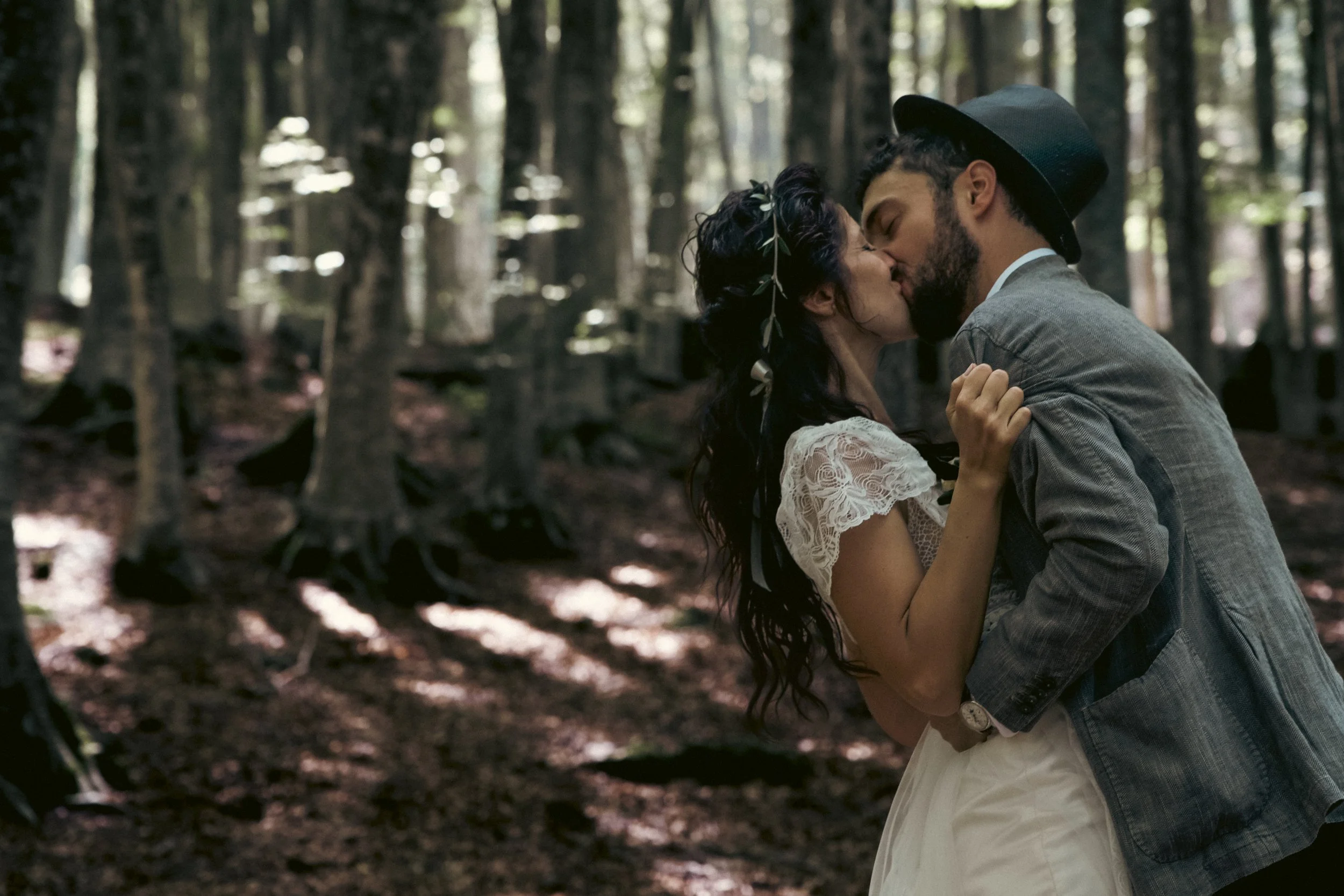 A couple sharing a kiss in a forest, woman wearing a white lace dress with leaf decorations in her hair, man wearing a gray jacket, black hat, surrounded by trees and sunlight.