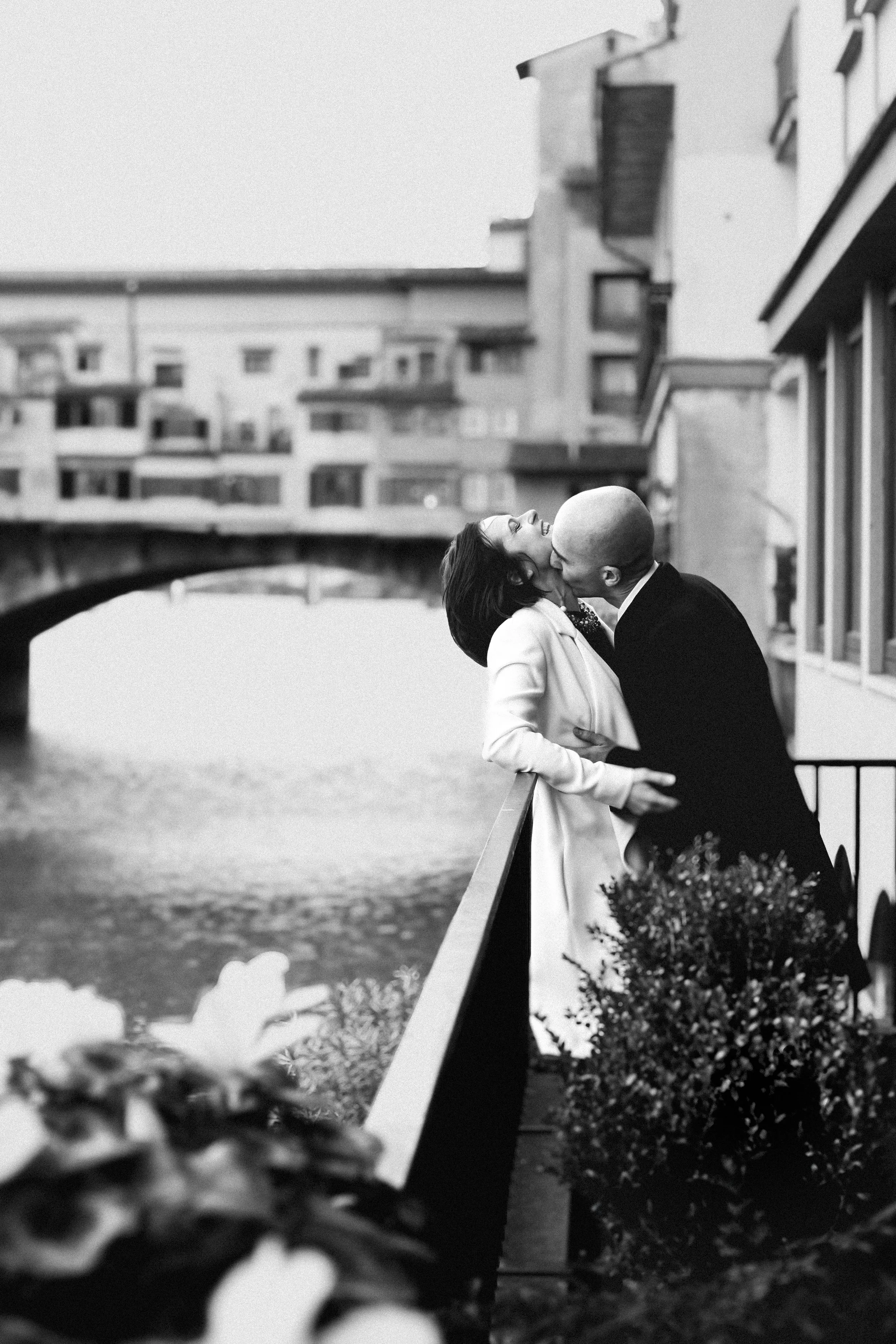 A black-and-white photo of a couple kissing on a balcony by a river, with an arched bridge and apartment buildings in the background.