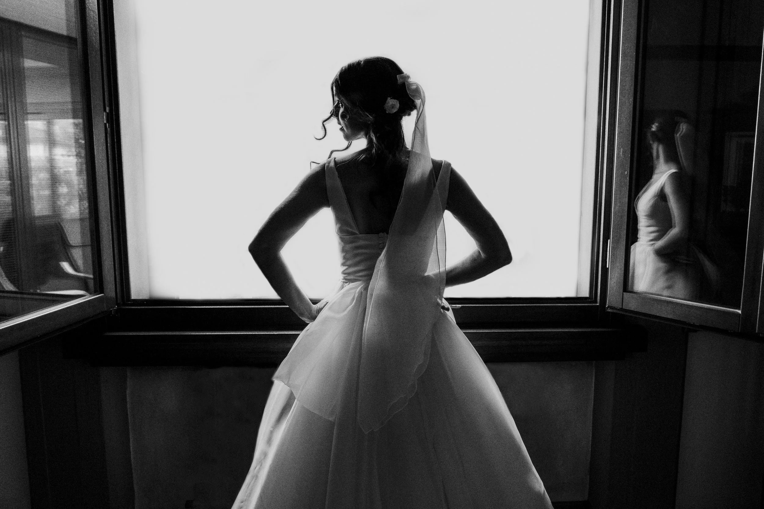 A woman in a wedding dress stands with hands on hips near a large window, looking away, with her reflection visible in a mirror on the right.