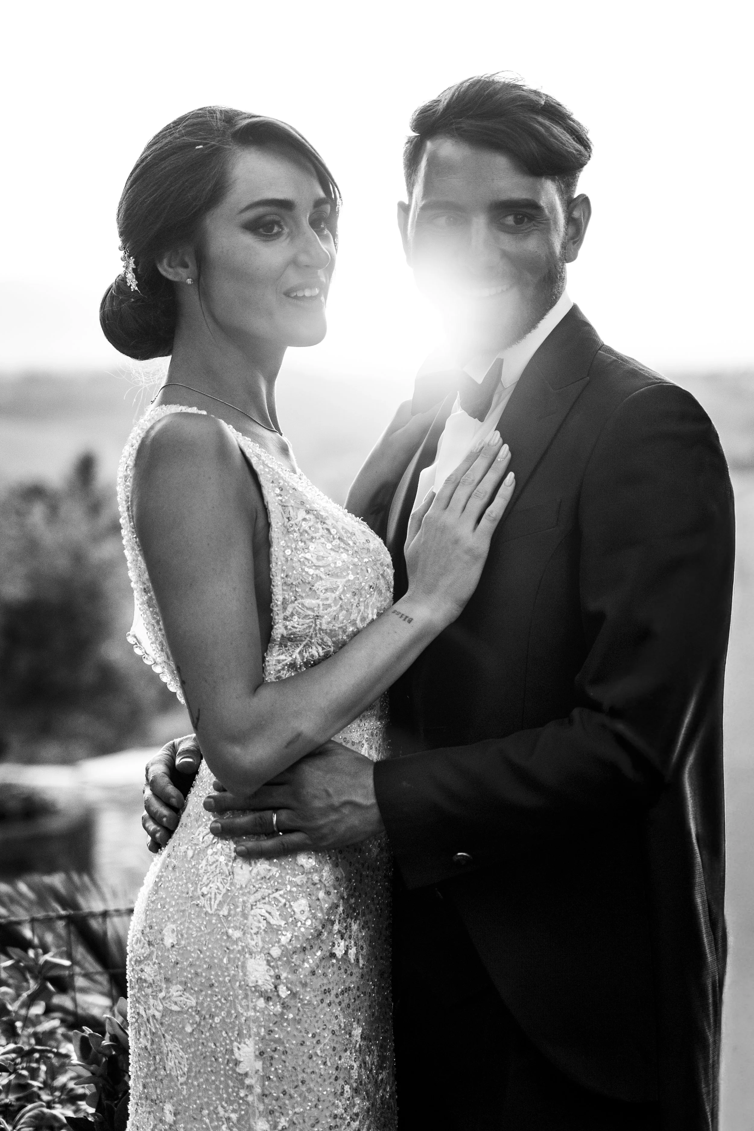 Black and white photo of a couple in wedding attire, smiling, with the bride's hand on the groom's chest, outdoors at sunset.
