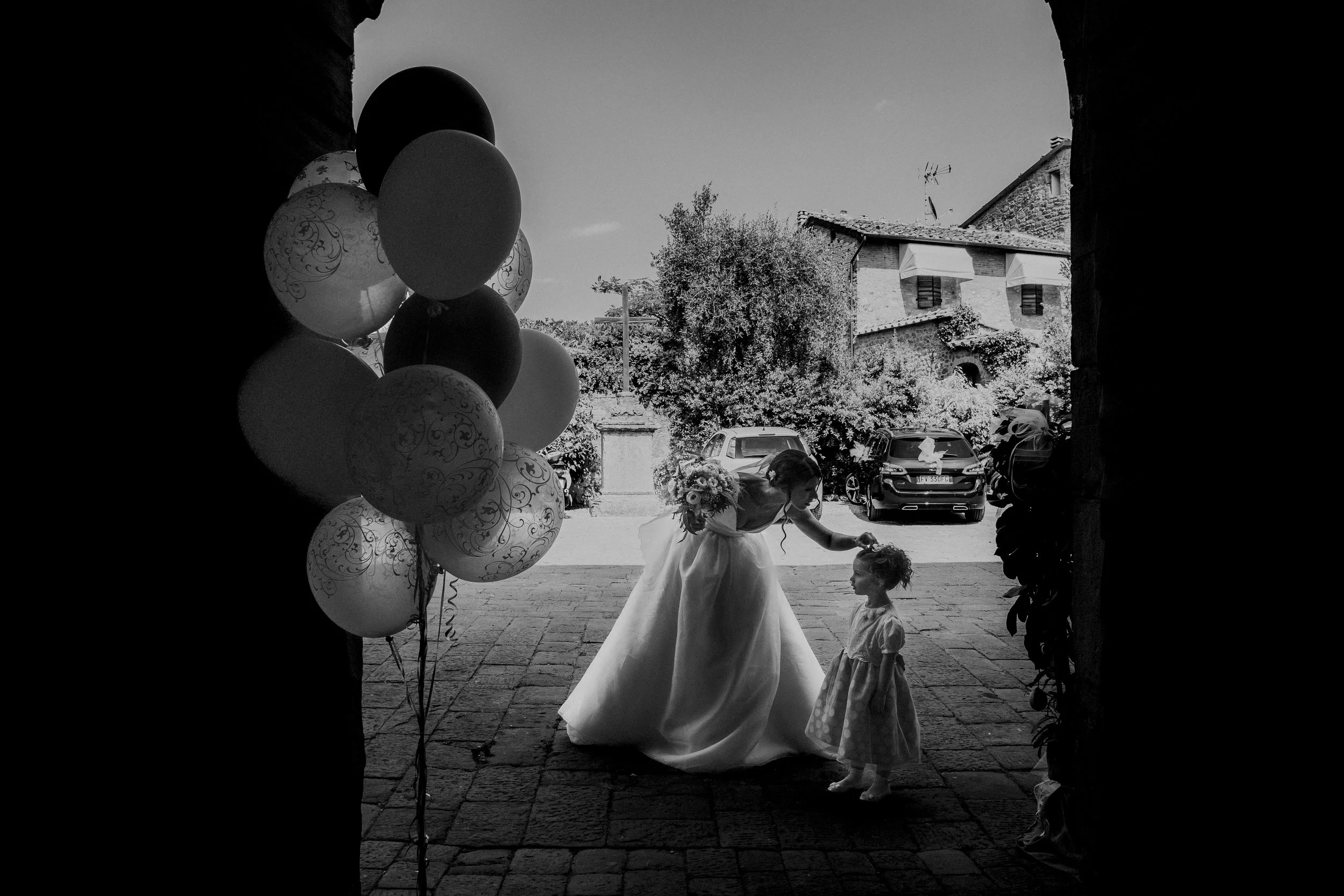 A woman in a wedding dress holding a bouquet and bending down to touch a young girl’s head, who is wearing a dress, viewed through an archway. Balloons are visible on the left side in the foreground, and cars and houses are in the background.