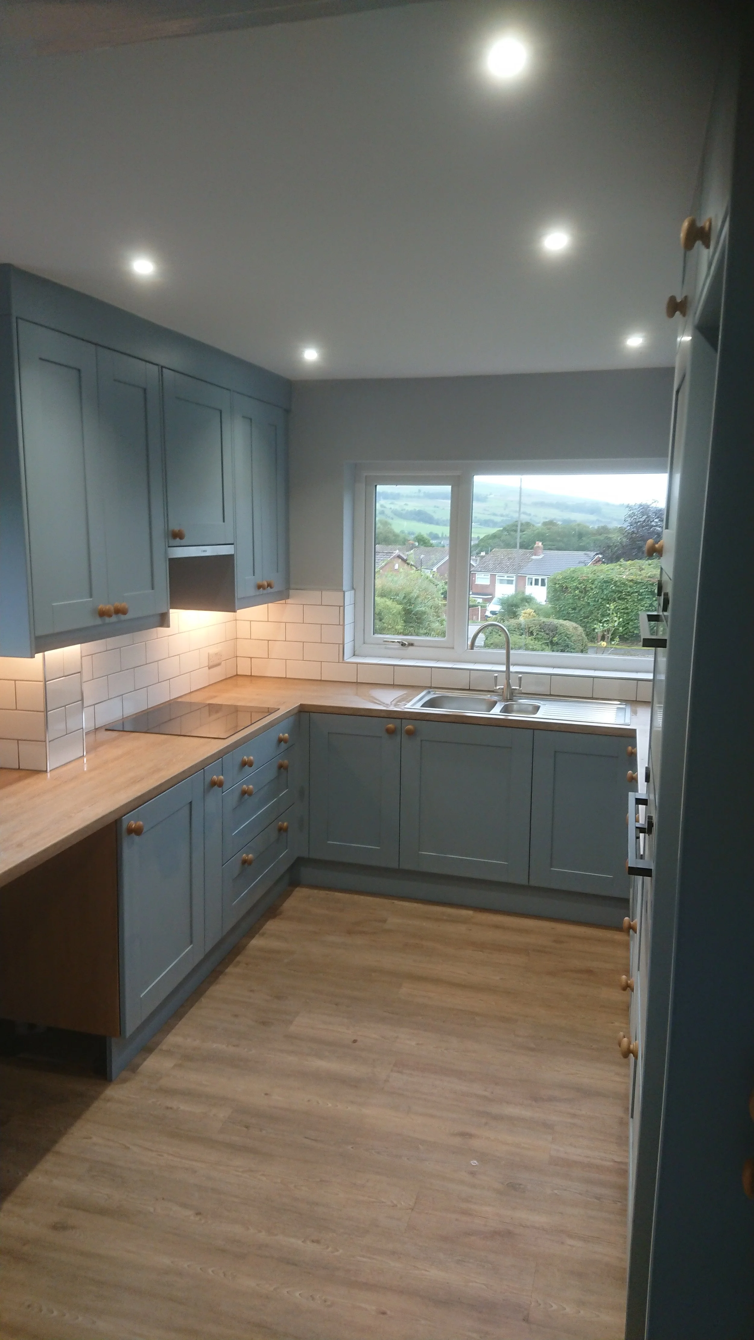 Modern kitchen with blue cabinets, wooden countertops, white tile backsplash, and a window overlooking a neighborhood with greenery.