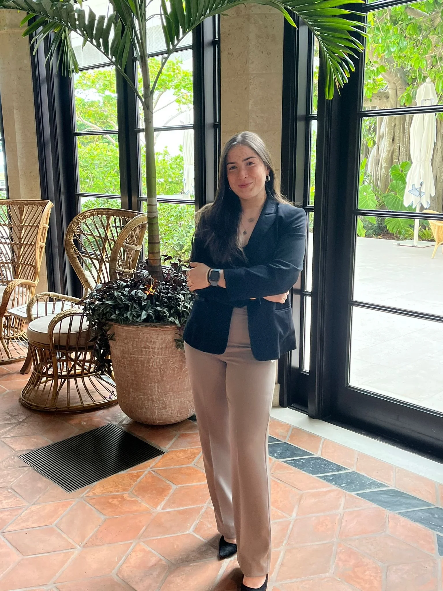 A woman with long dark hair wearing a black blazer and beige pants standing inside a sunlit room near large glass doors. There are wicker chairs and a large potted plant nearby, with green foliage visible outside.